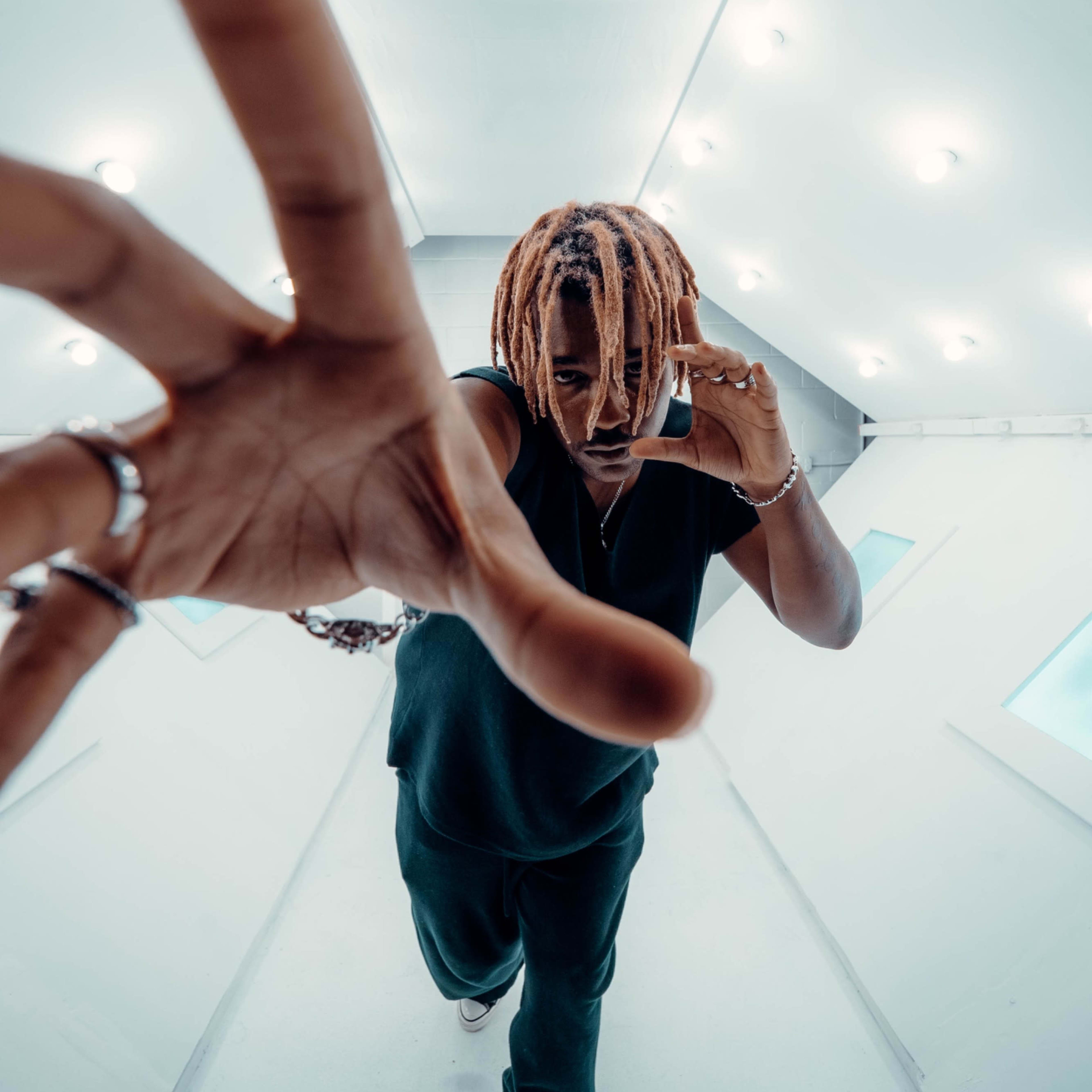 A person with dreadlocks holds a phone to their ear while extending one hand toward the camera in a white, brightly lit space.