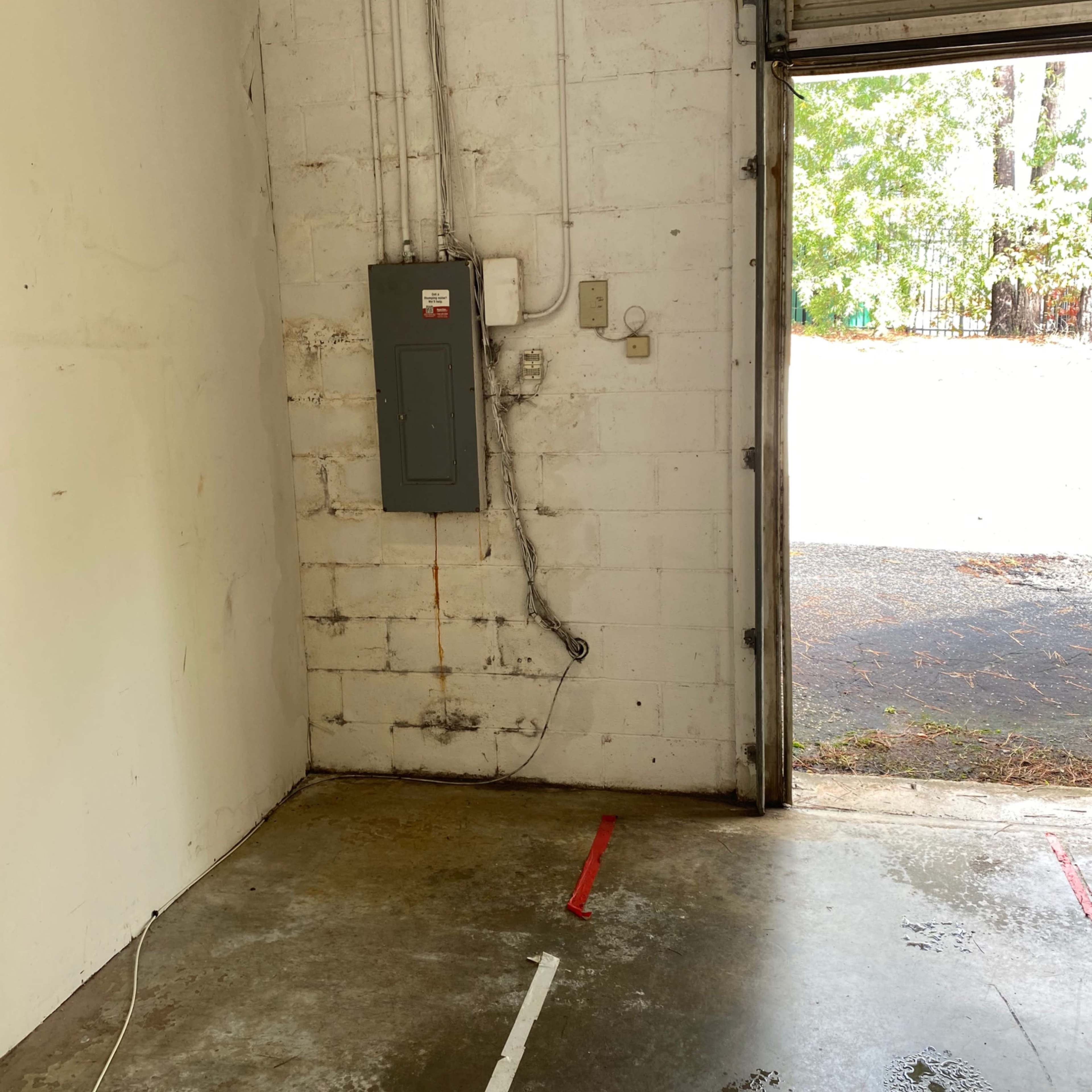 A partially empty garage with a concrete floor, a utility panel on the wall, and an open roll-up door leading to an outdoor area.