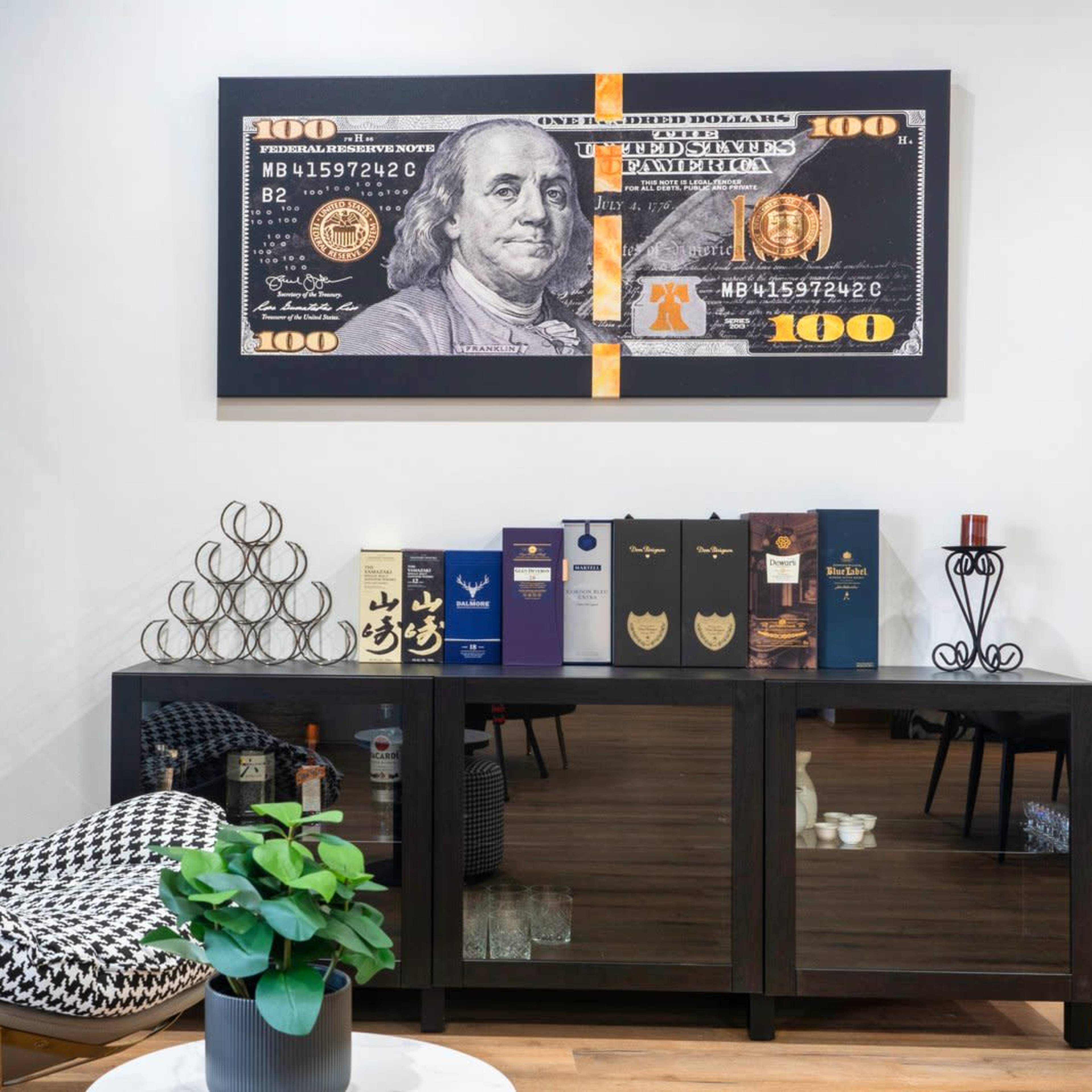 A modern interior showcases a black and white patterned chair next to a sleek cabinet displaying decorative items and a large framed image of a one hundred dollar bill on the wall.
