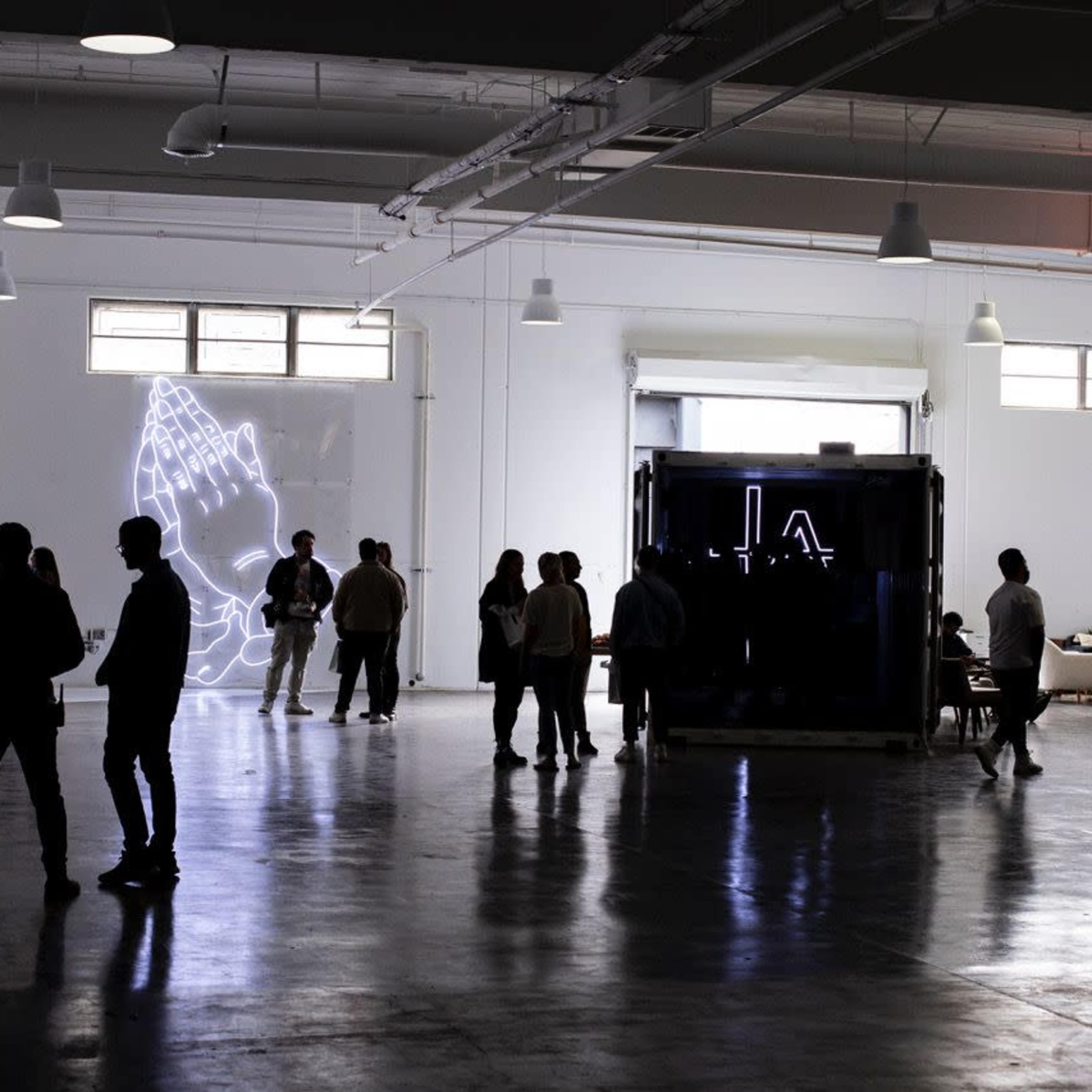 A spacious indoor venue with a large neon artwork of clasped hands on the wall and several groups of people chatting in the foreground.