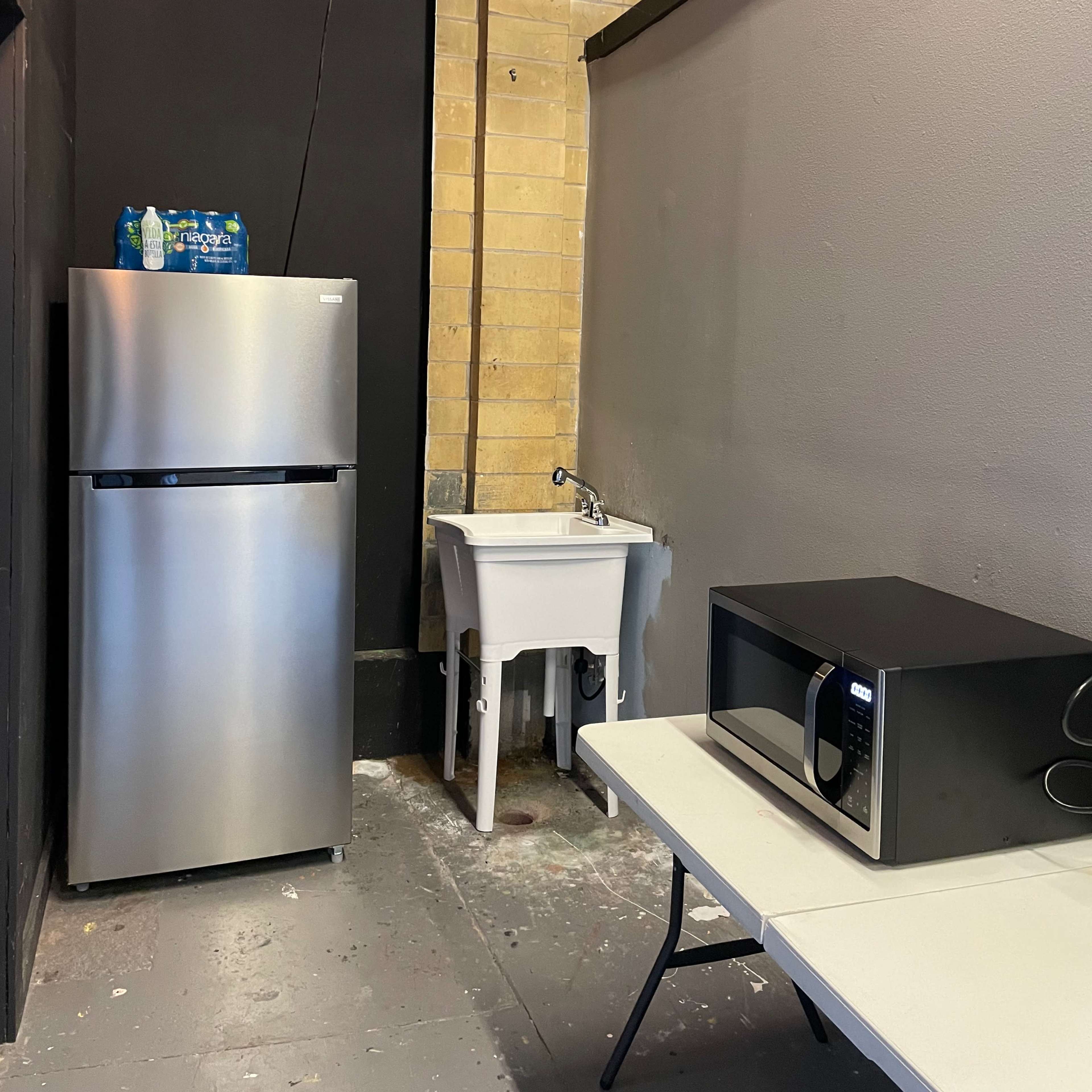 A small kitchenette area featuring a silver refrigerator, a microwave on a table, and a sink against a wall with exposed brick.