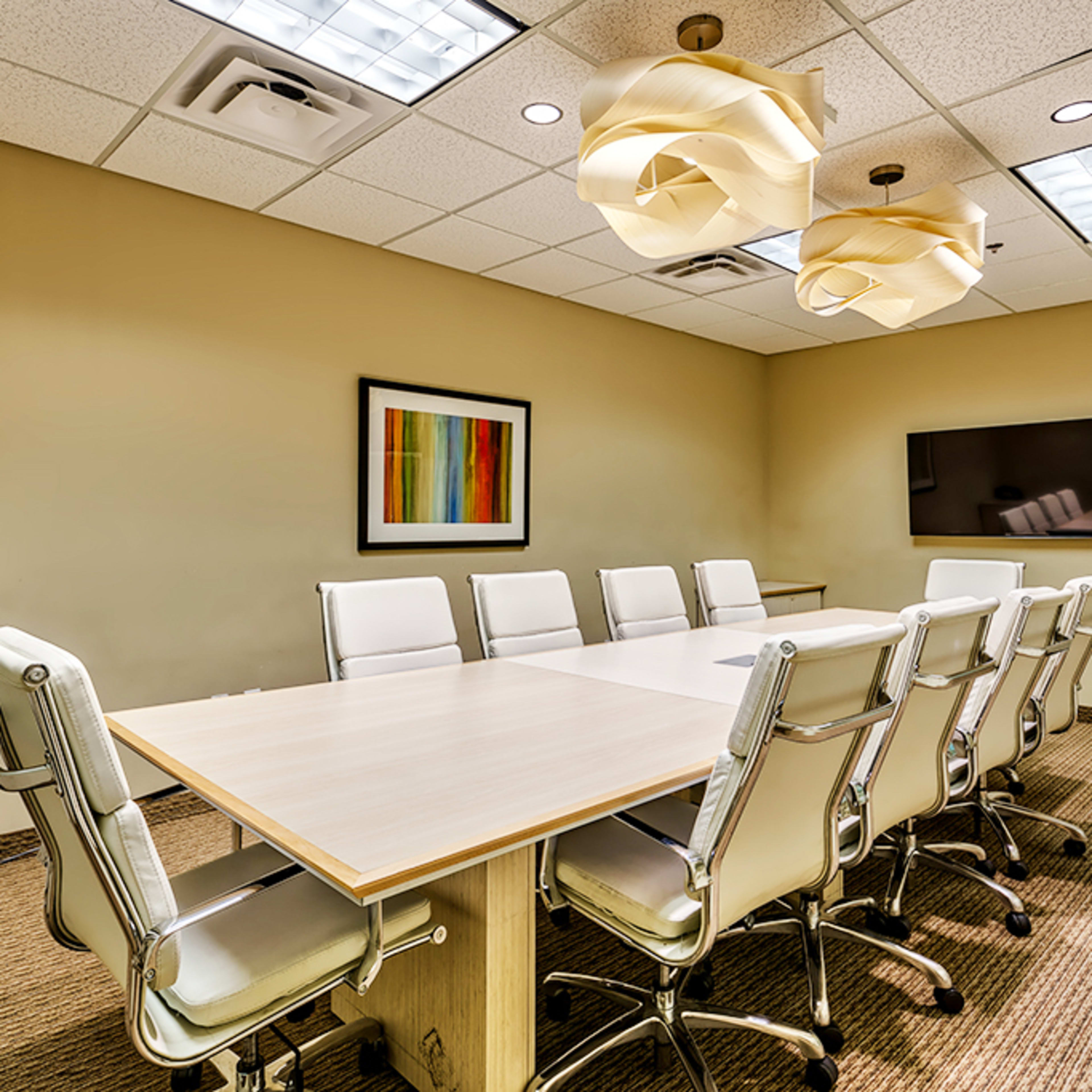 A modern conference room features a long table surrounded by white chairs, with decorative light fixtures on the ceiling and a television mounted on the wall.