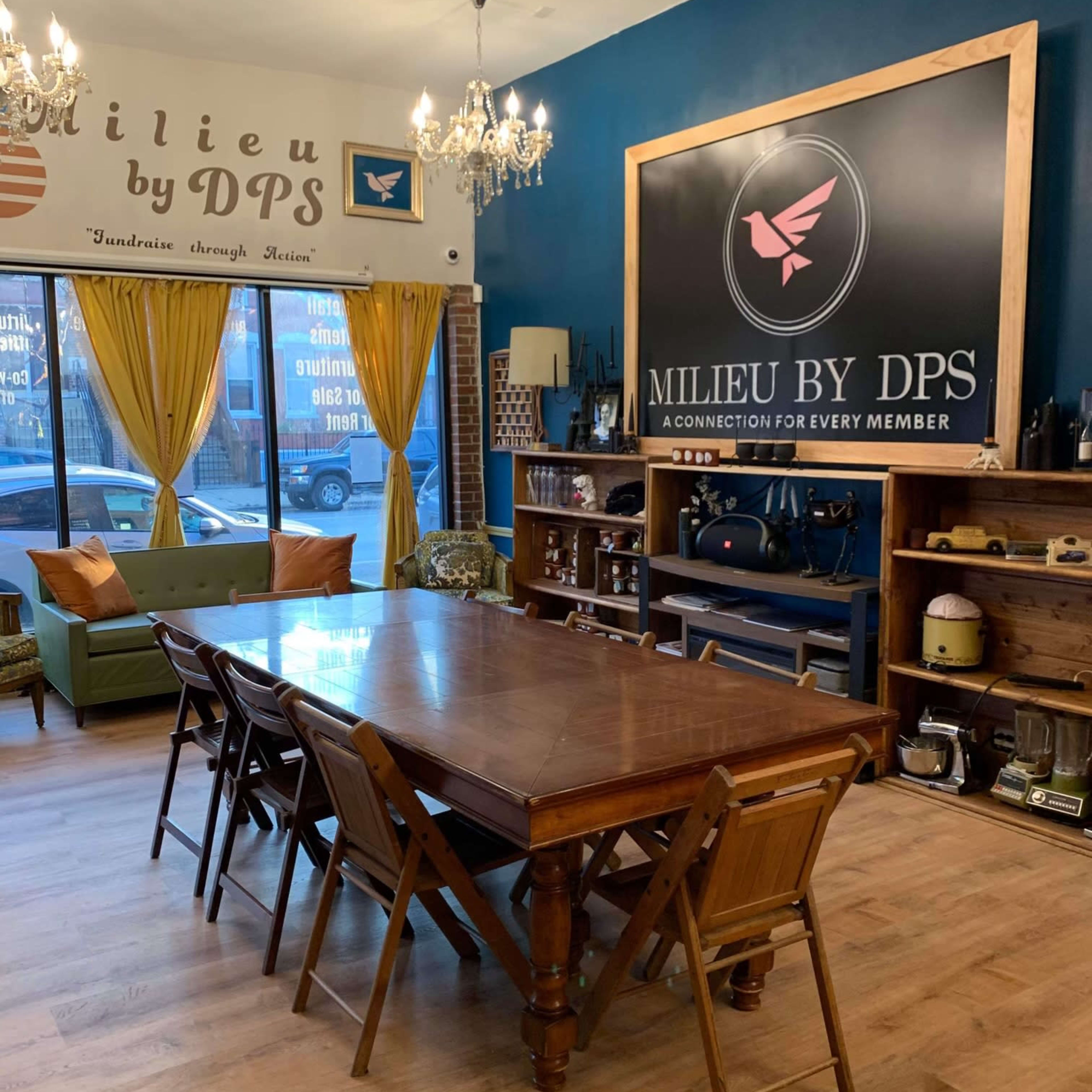 A well-lit meeting space featuring a large wooden table surrounded by folding chairs, with a sign for "Milieu by DPS" on the wall and large windows draped with yellow curtains.