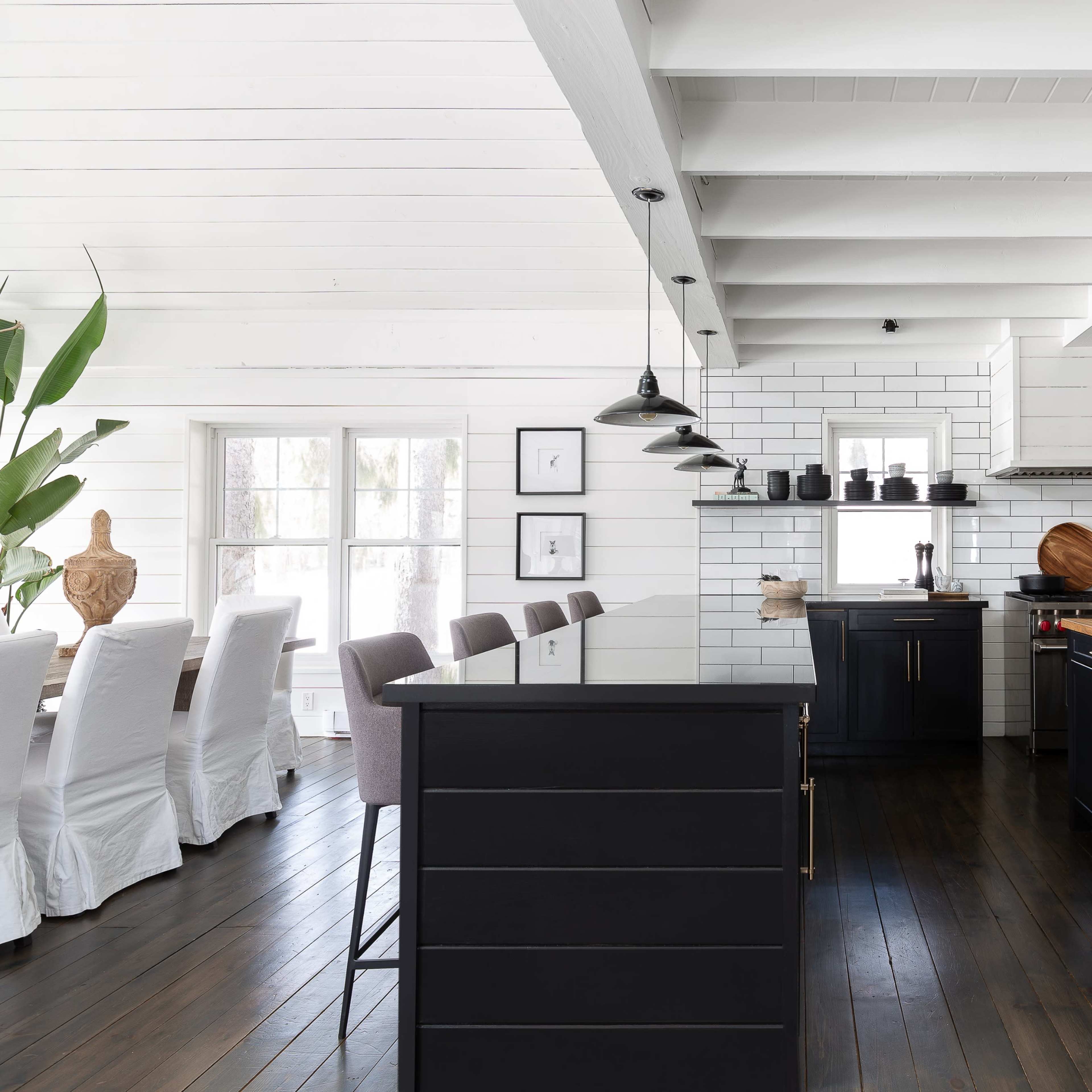 A modern kitchen features black cabinetry, a wooden countertop, and a white-tiled backsplash, with an adjoining dining area that has a long table and light-colored chairs.
