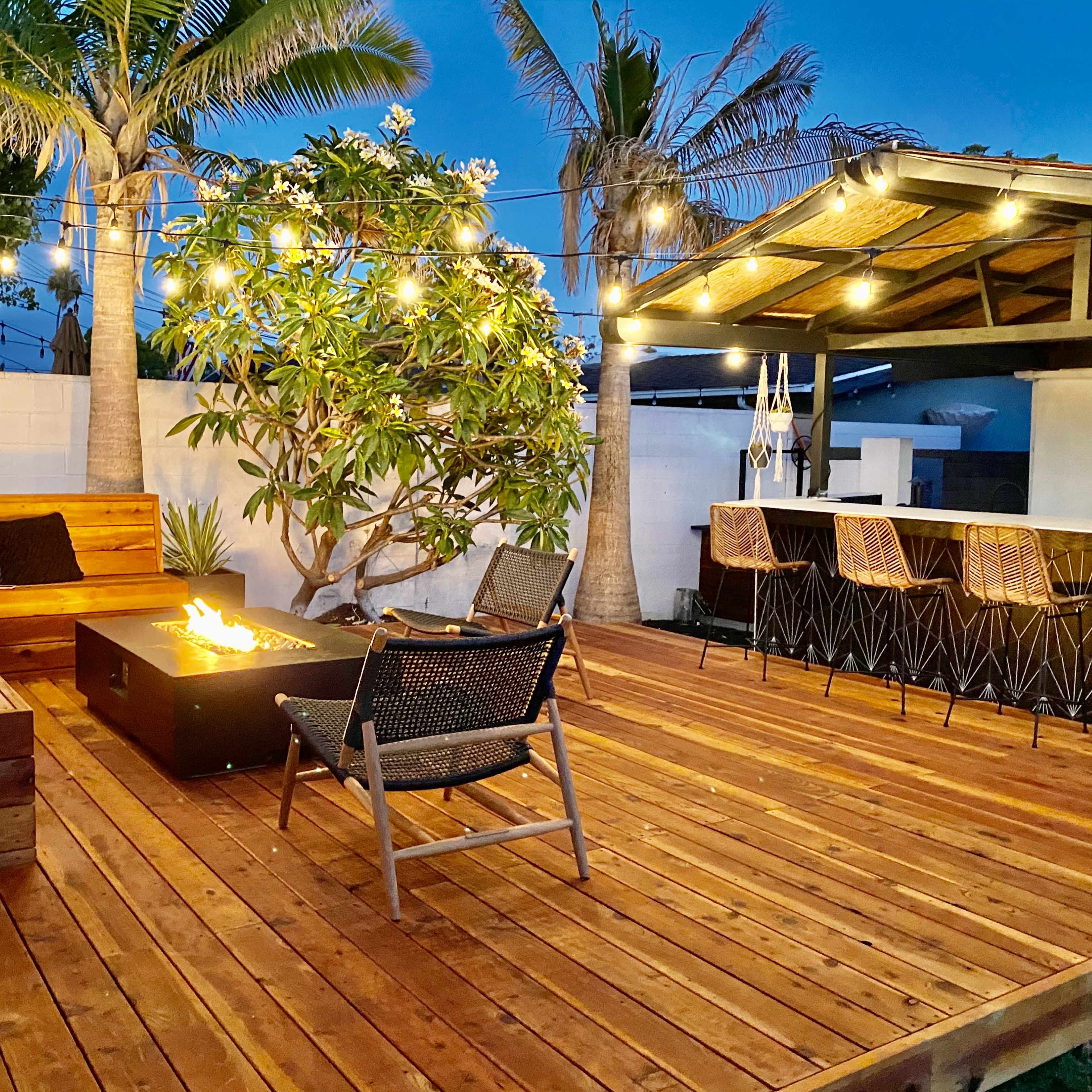 A wooden deck with a fire pit surrounded by outdoor seating, illuminated by string lights in the evening.