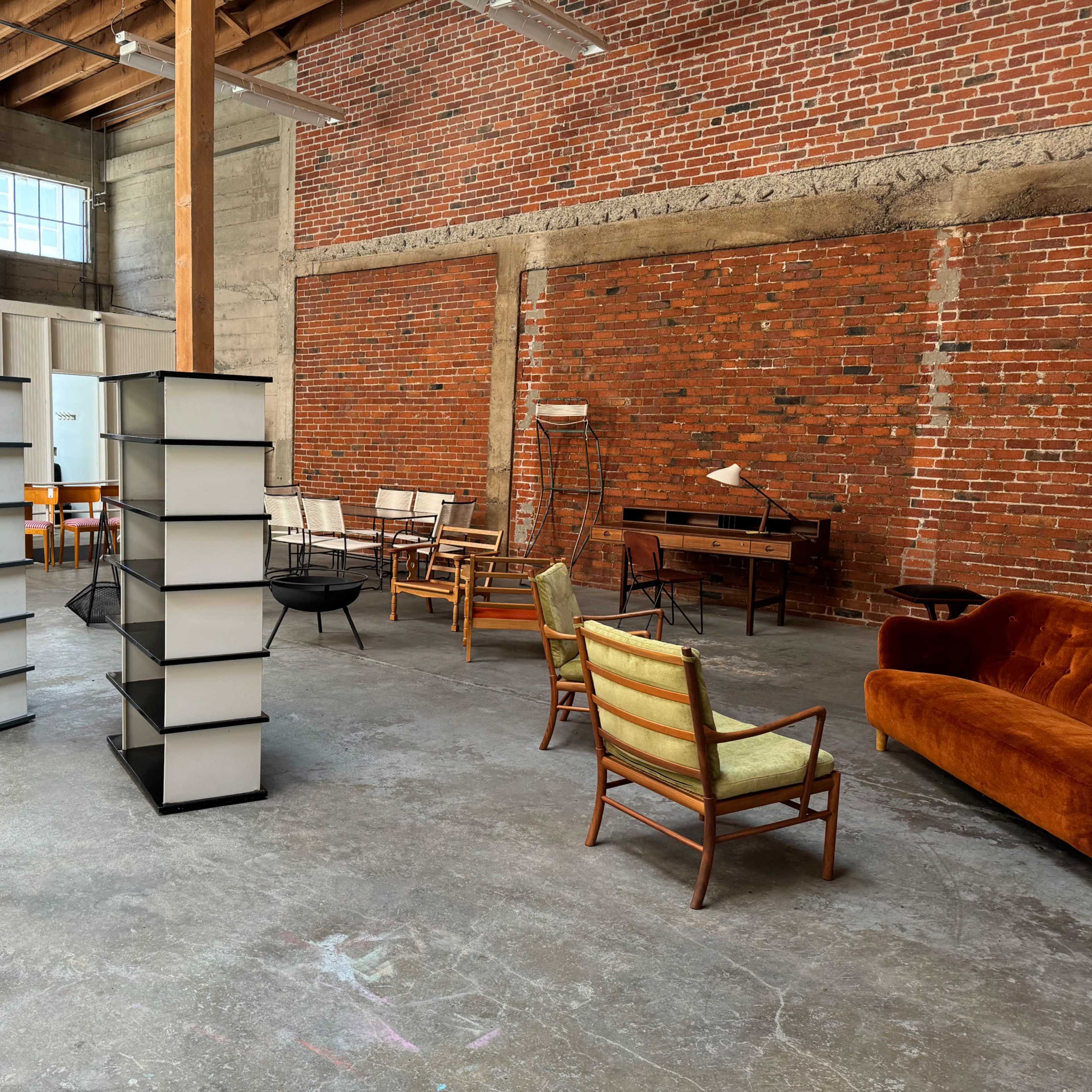 An open, industrial-style interior featuring shelving units, various chairs, and a desk arranged on a concrete floor against a backdrop of exposed brick walls.