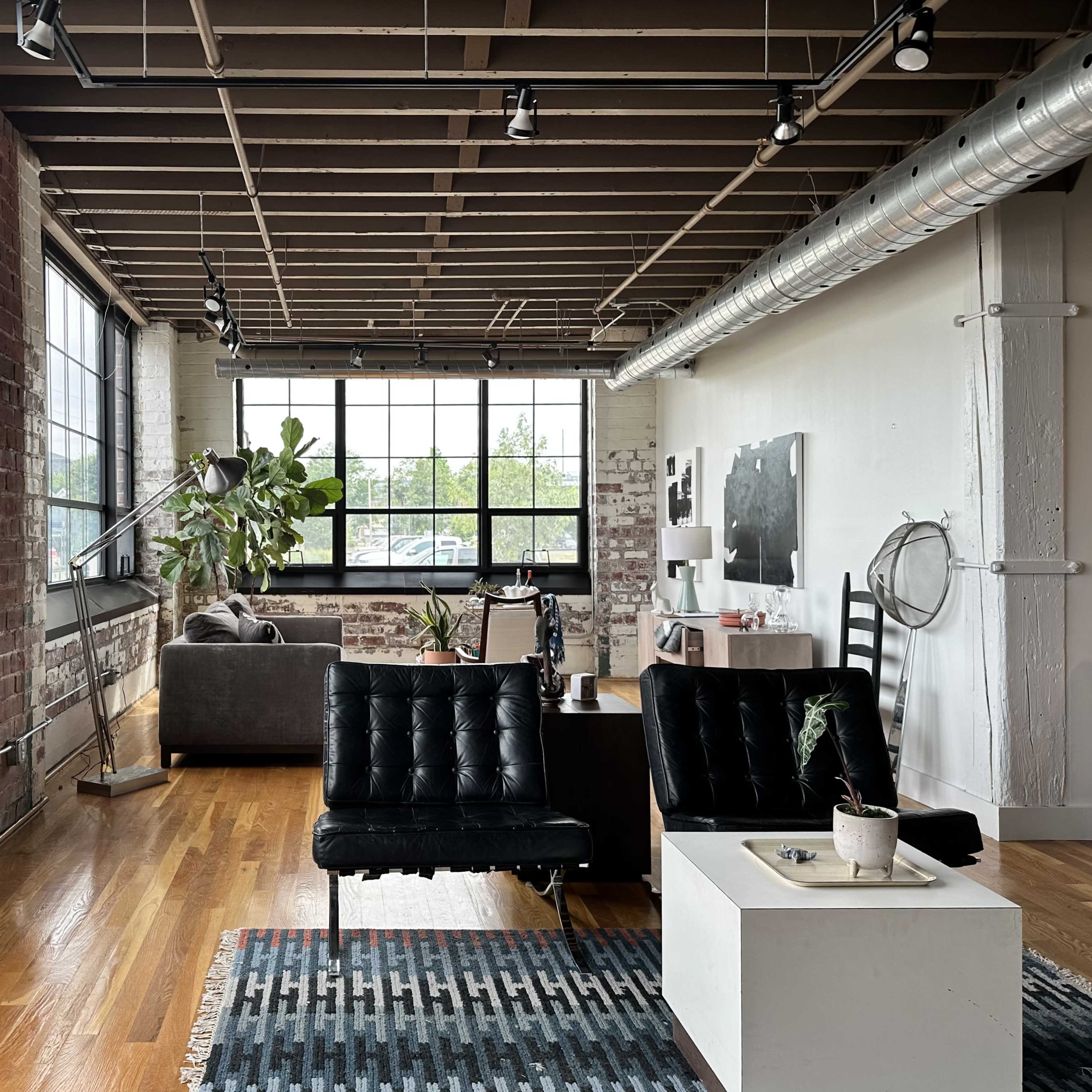 A modern loft features exposed brick walls, large windows, and a mix of contemporary furniture, including a black leather couch and a white coffee table.