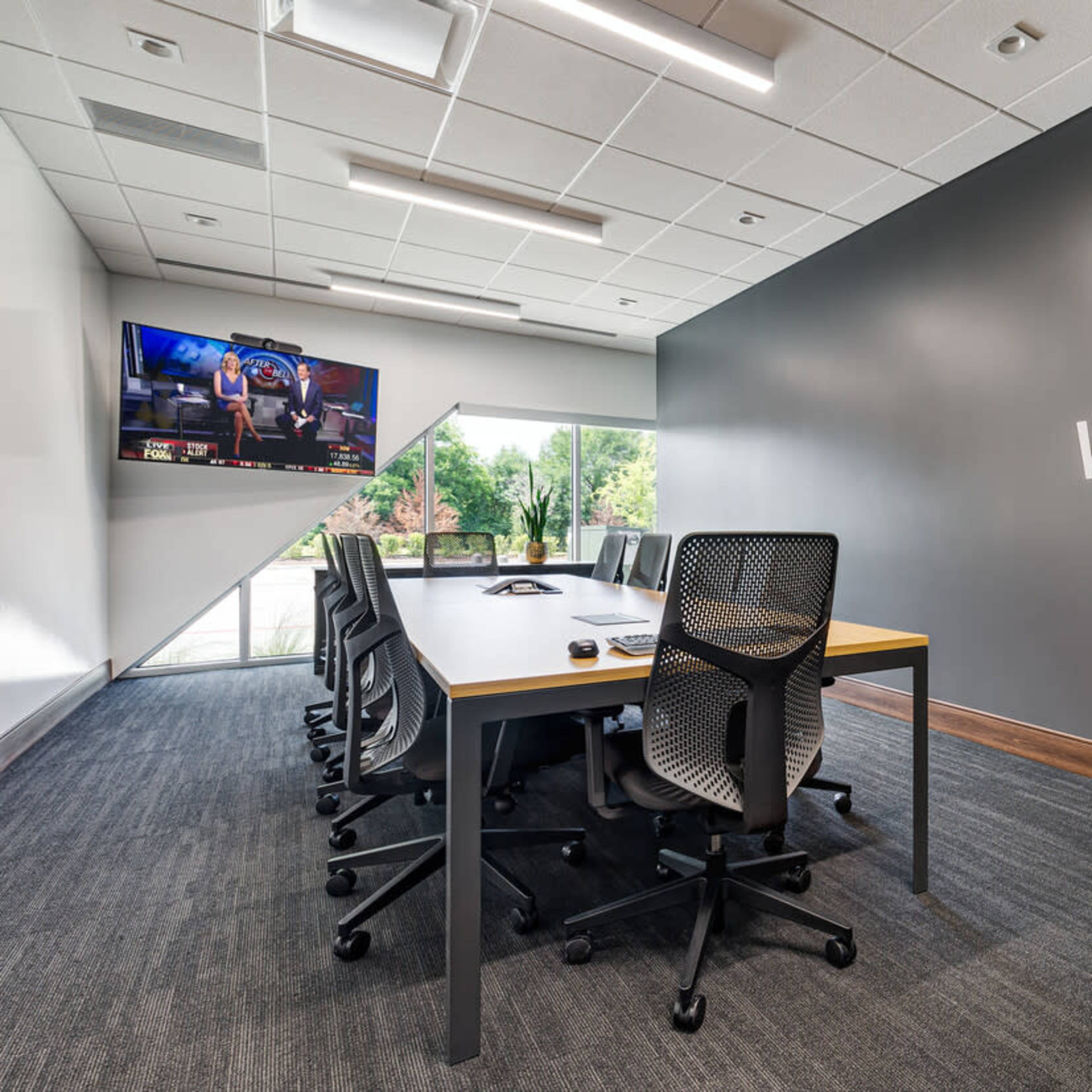 A modern conference room features a long table surrounded by ergonomic chairs, with a television mounted on the wall and a large window providing natural light.