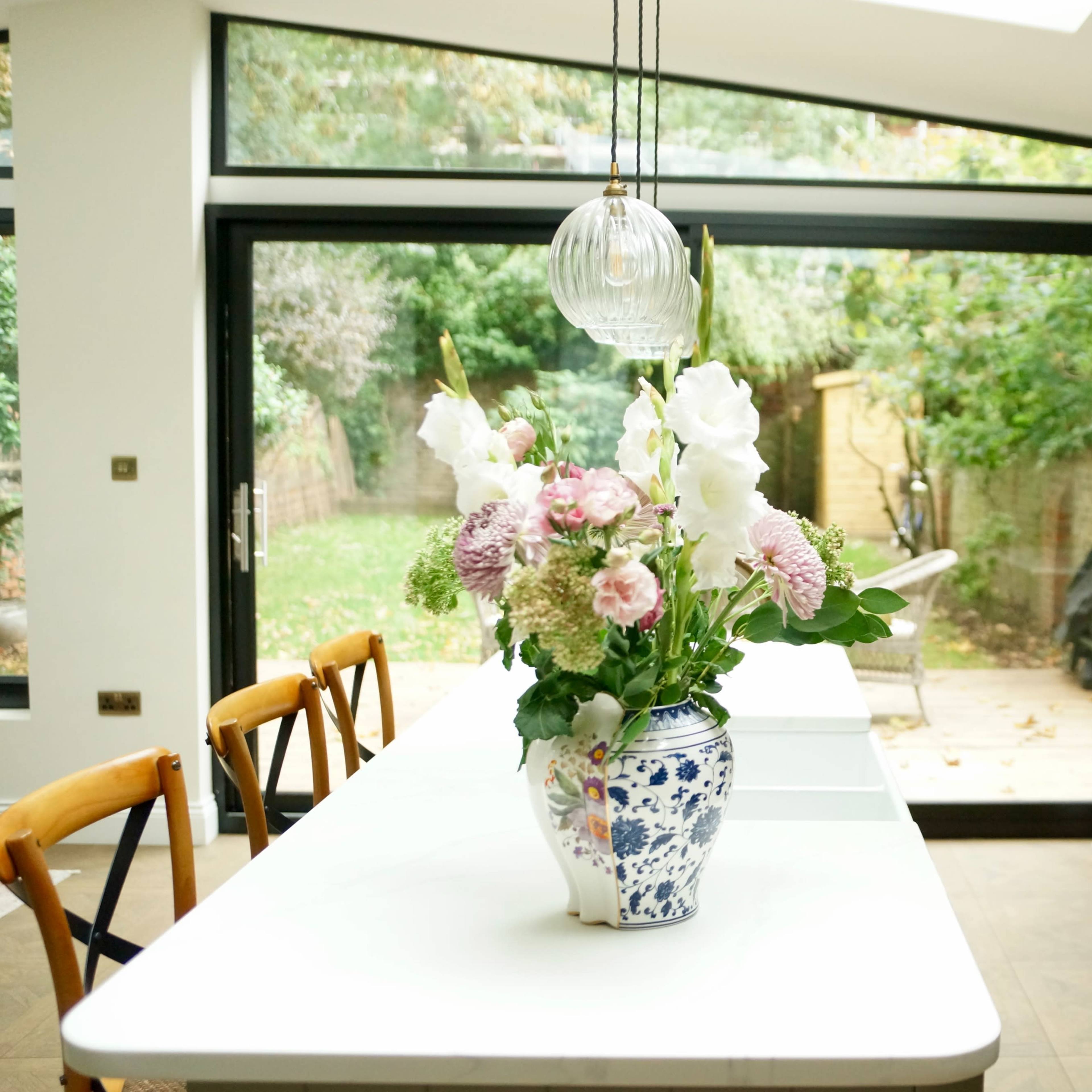 A modern kitchen features a large island with a floral arrangement in a vase, and large glass doors that open to a garden.