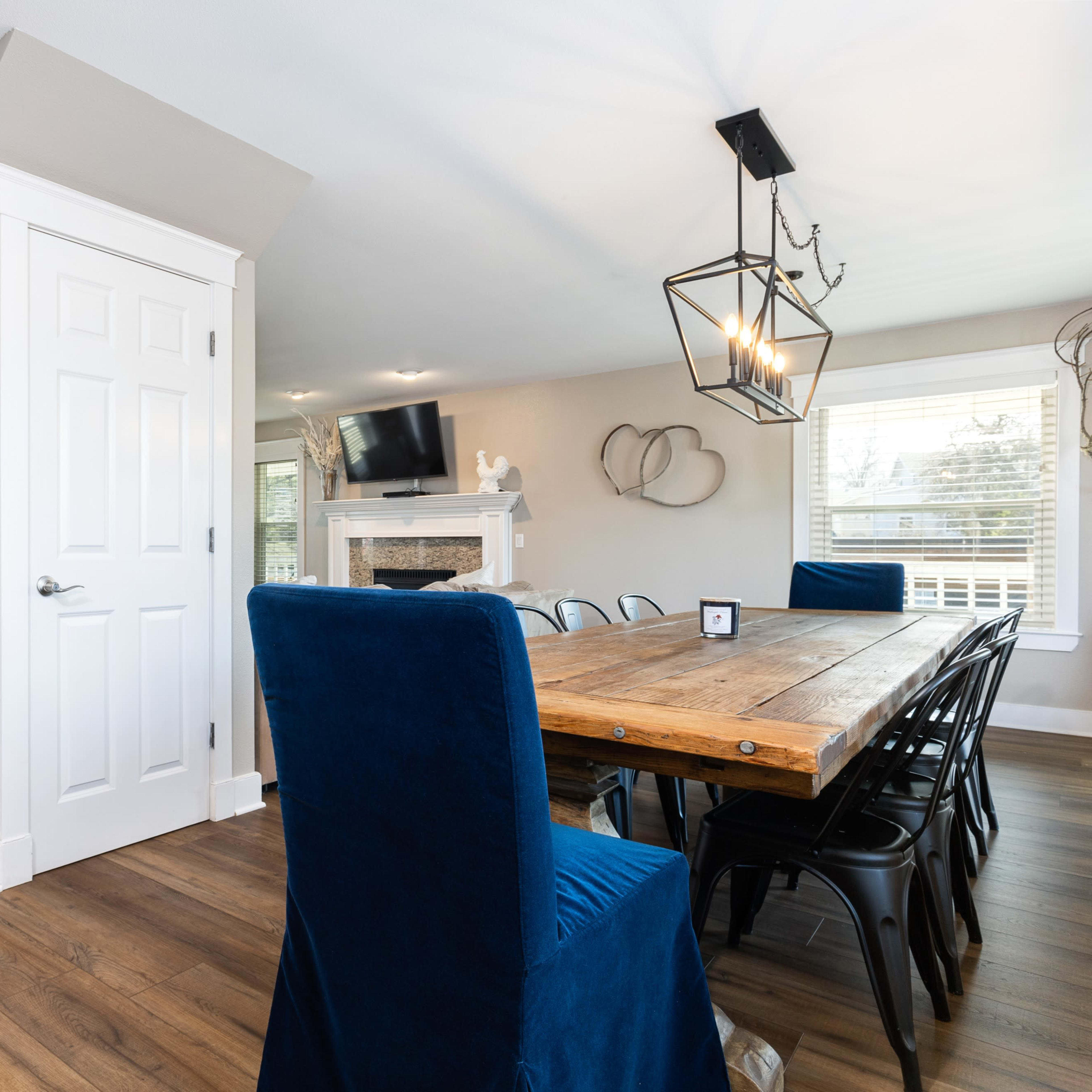 A modern dining area with a wooden table surrounded by blue chairs, leading to a staircase and an open living space with a fireplace and television.