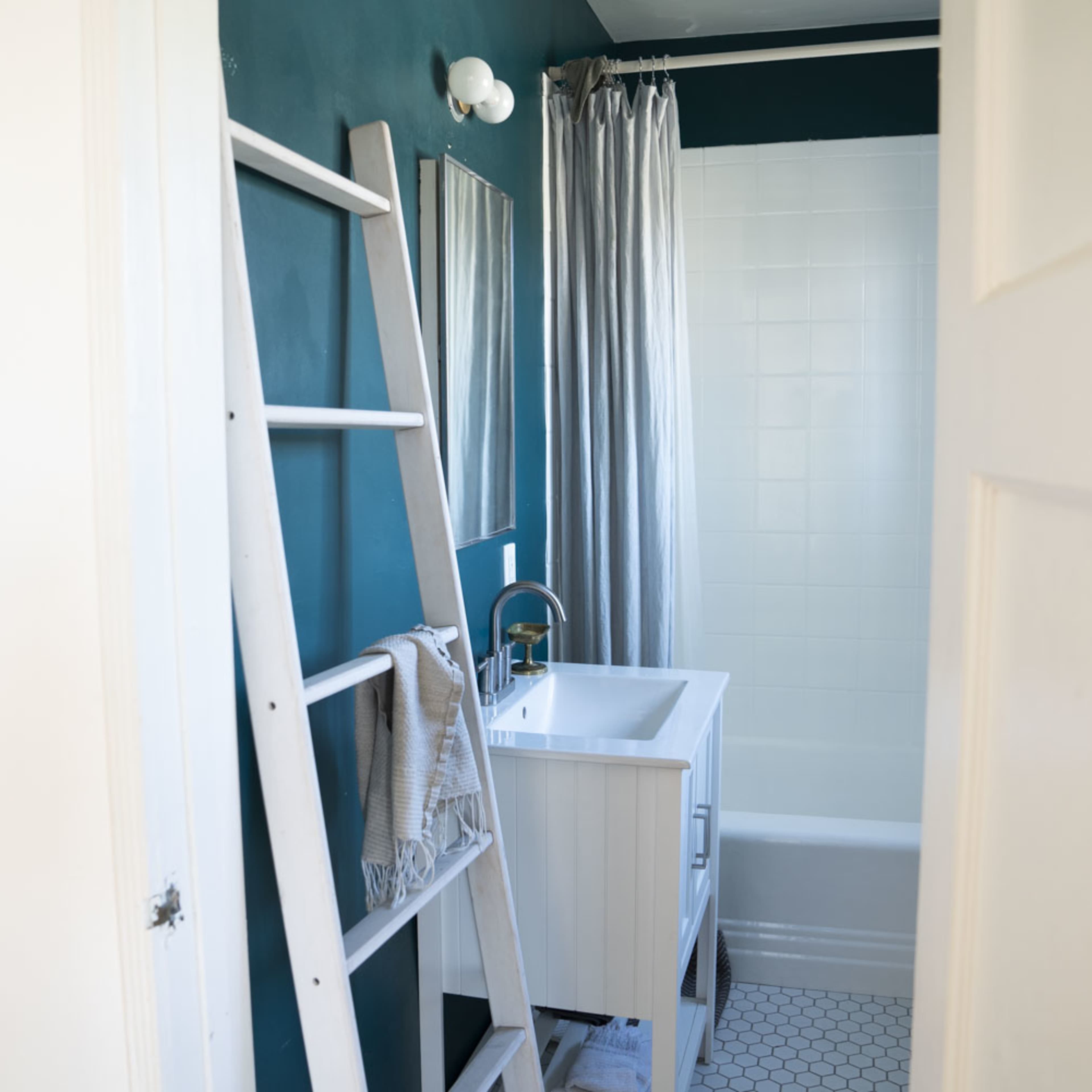 A bathroom features a tub, a white vanity with a sink, a mirror, and a shower curtain, with a ladder leaning against the wall.
