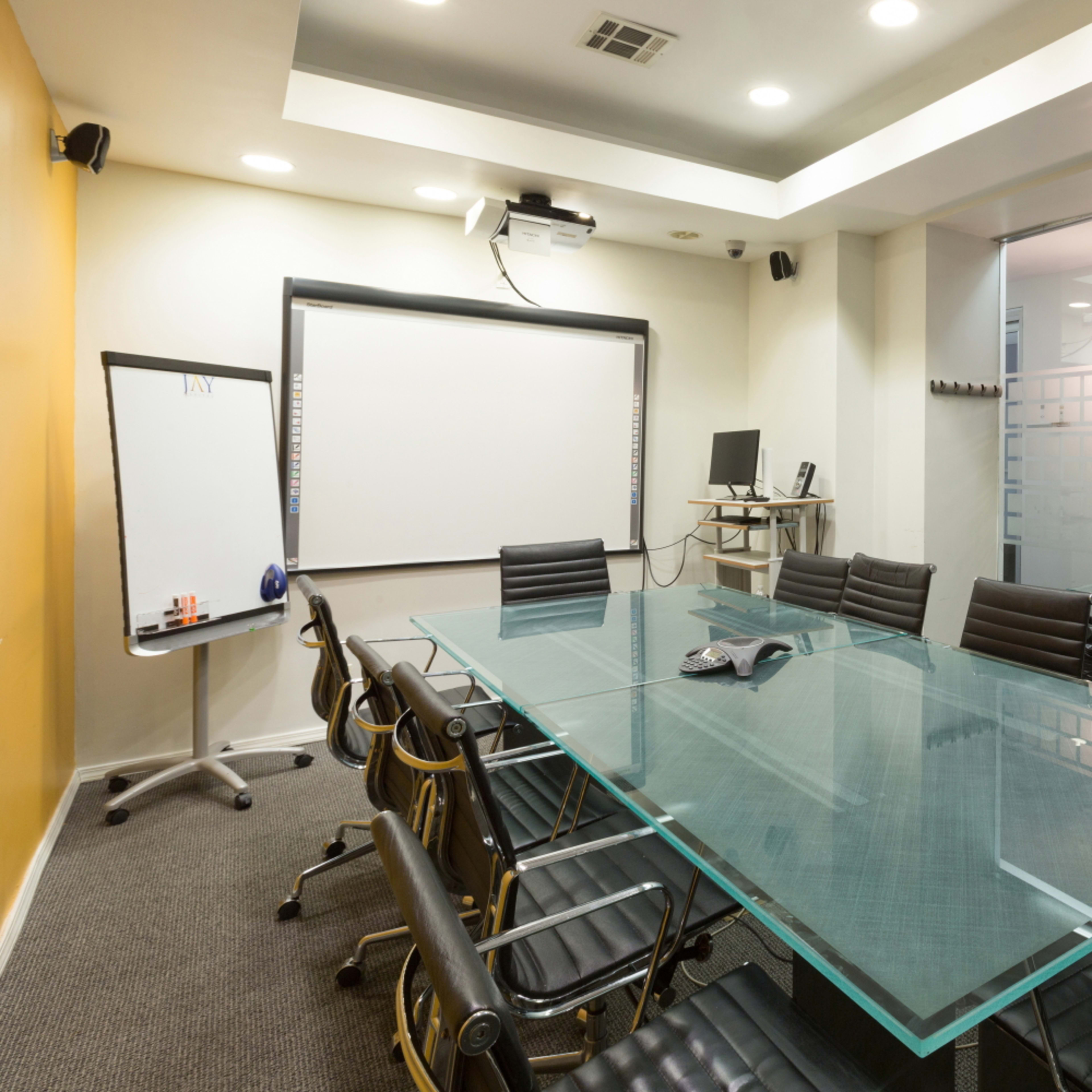 A modern conference room features a large glass table surrounded by black chairs, with a projector, whiteboard, and glass wall.