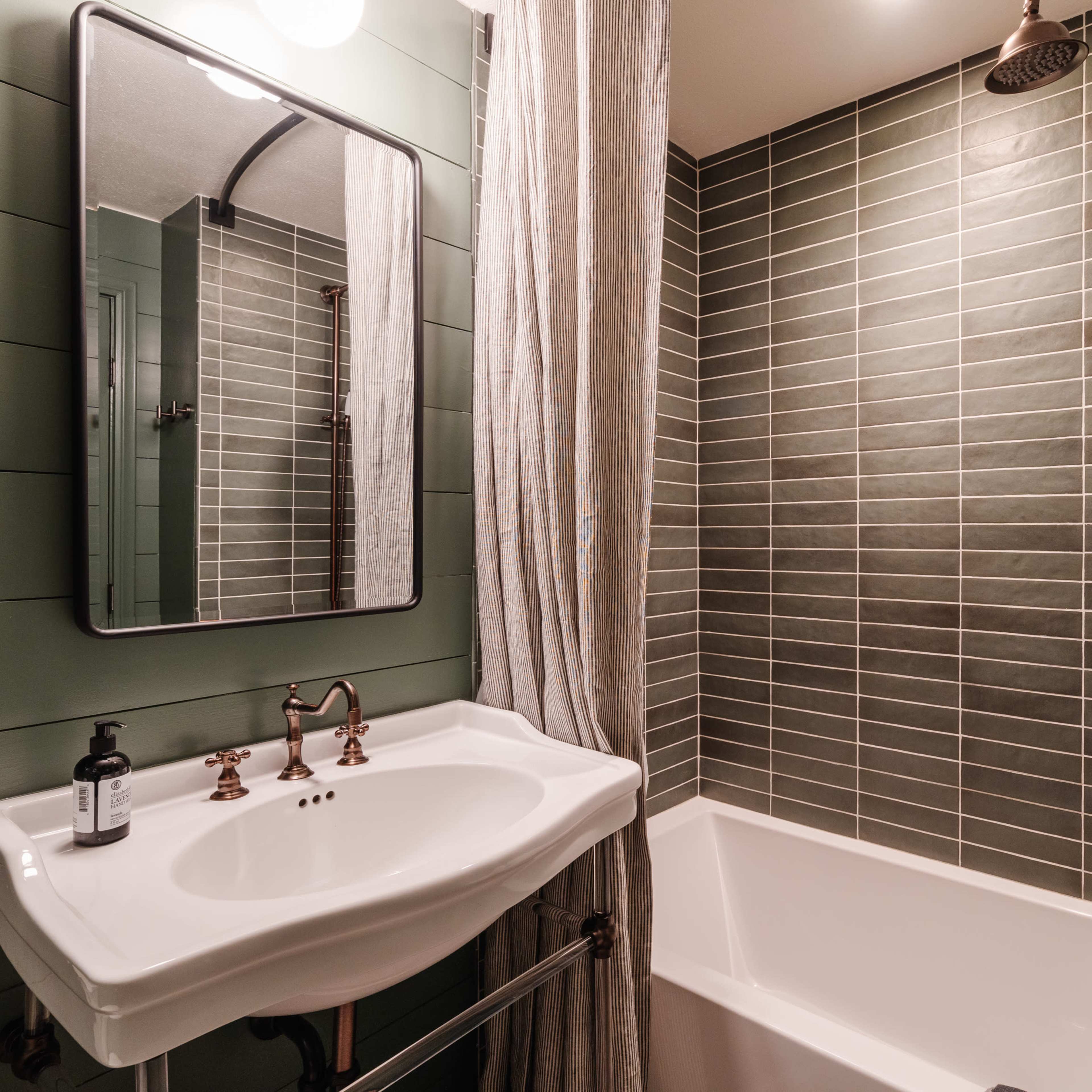 A bathroom with a green tiled wall, a white bathtub, a sink with brass fixtures, and a large mirror.