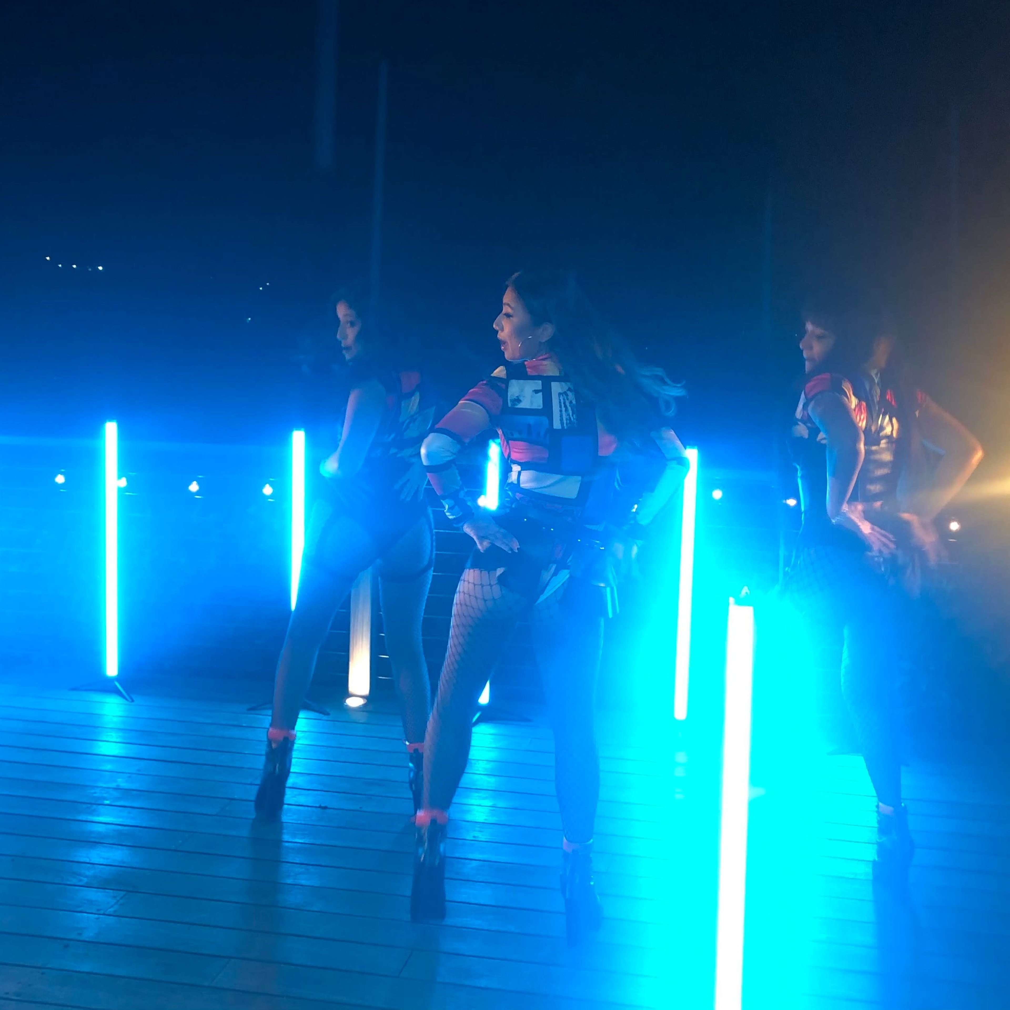 Three dancers in fitted outfits perform on a dimly lit stage with bright blue lighting.