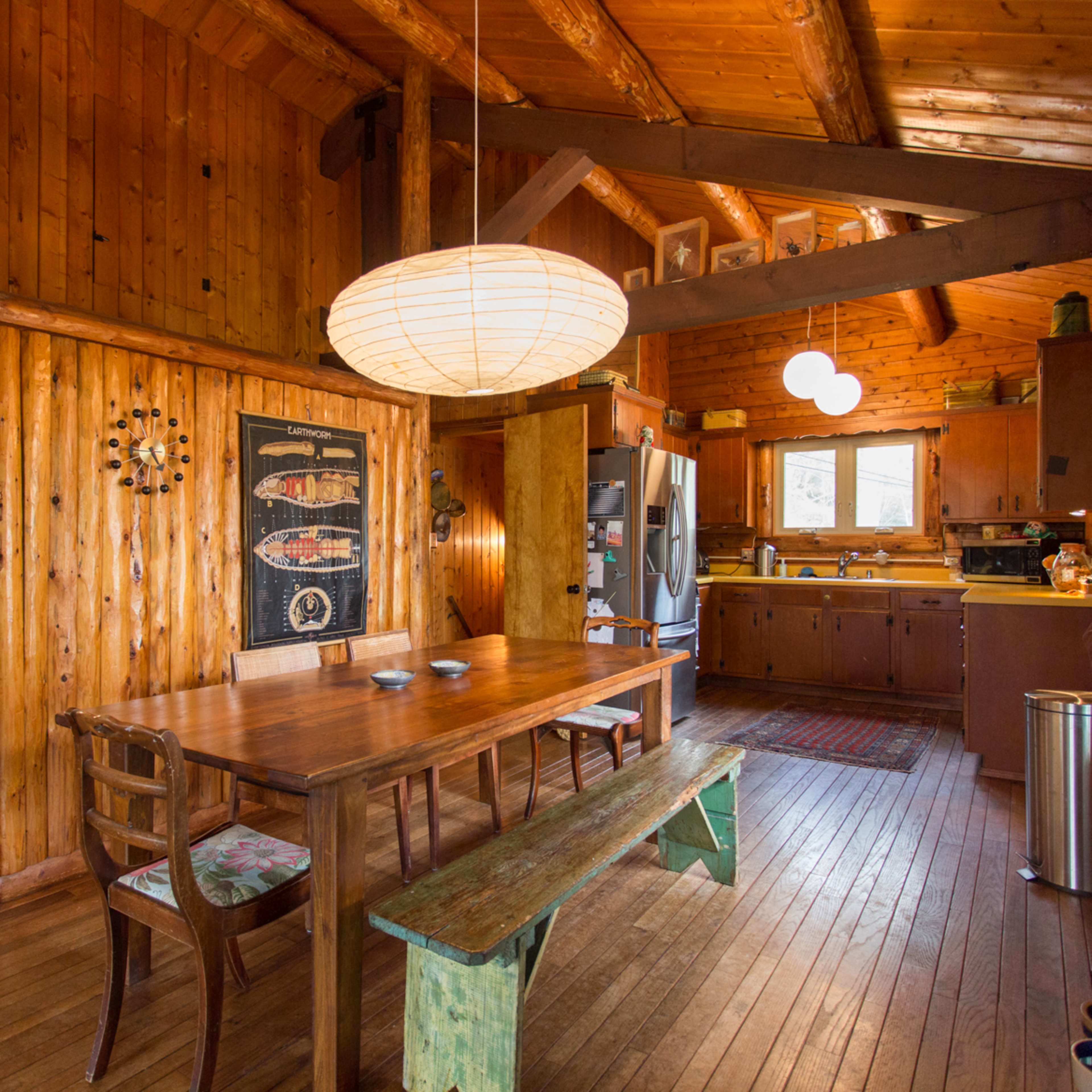The scene features a wooden dining area with a large table, surrounded by chairs, next to a kitchen with a window and hanging light fixtures.