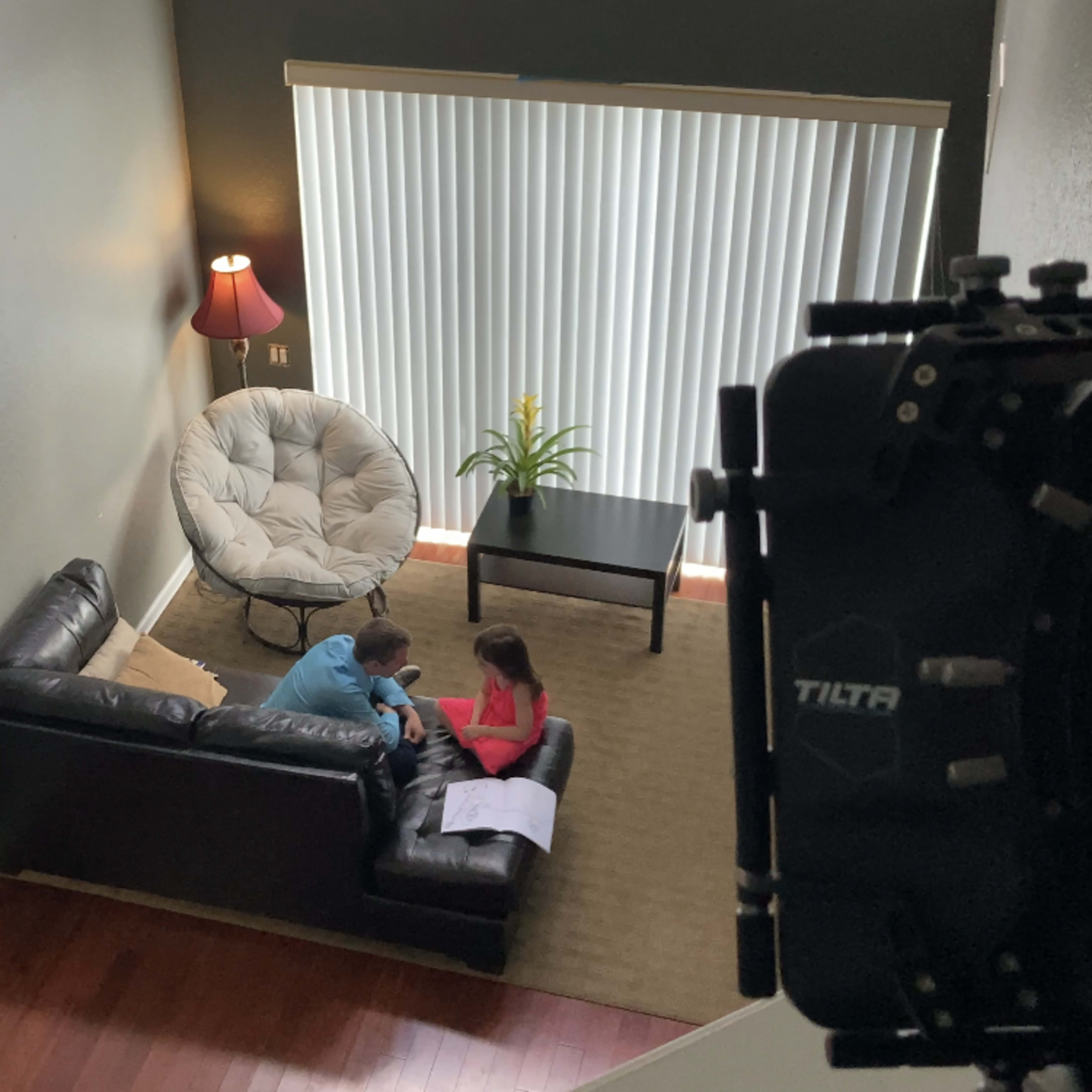 Two children sit on a black sofa in a living room, engaging with a paper on a coffee table, while a large floor lamp and a plant are nearby.