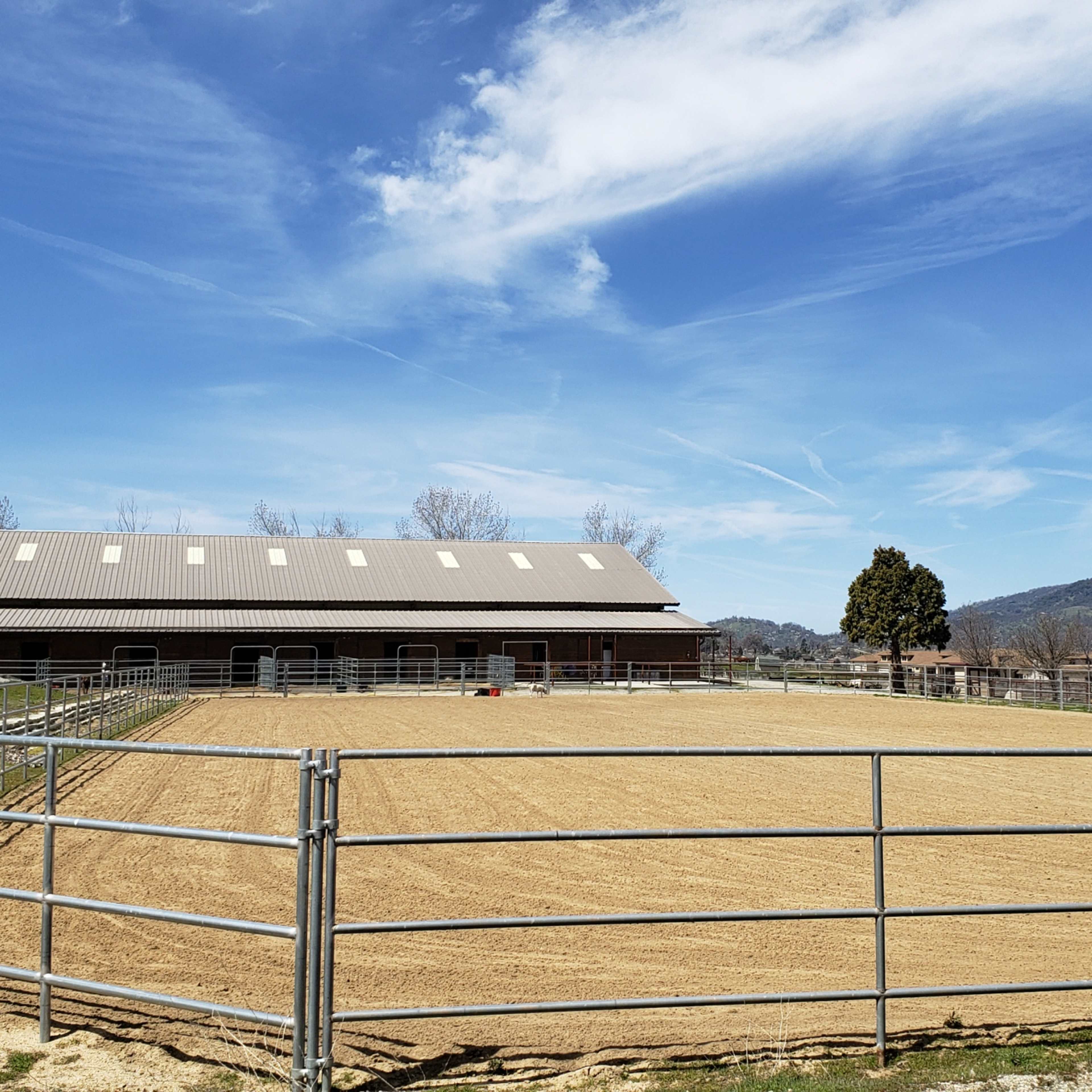 A ranch area with a fenced riding arena, a large barn, and a backdrop of rolling hills under a blue sky.