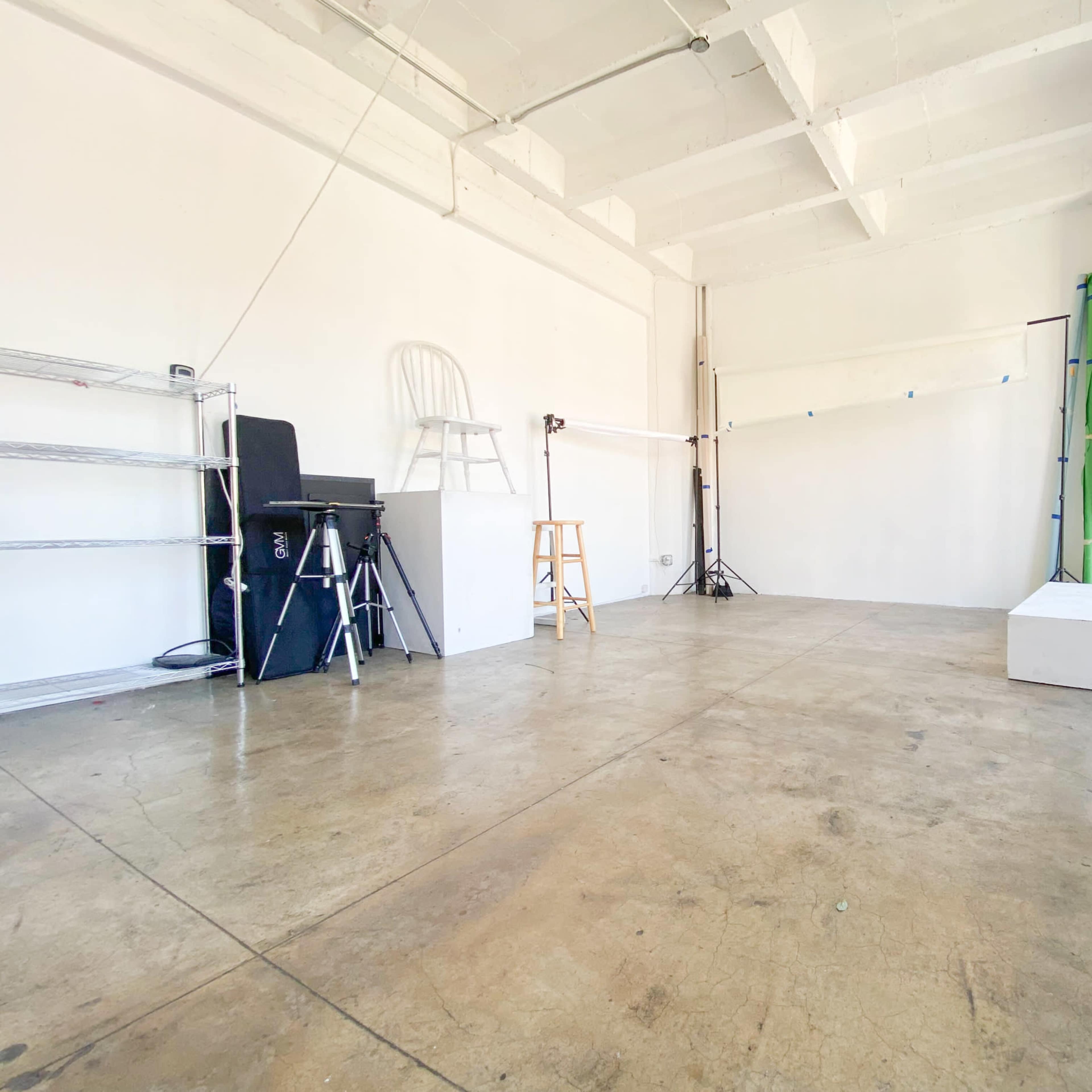 An empty studio space with a concrete floor, white walls, and various photography equipment visible, including a chair and a backdrop setup.