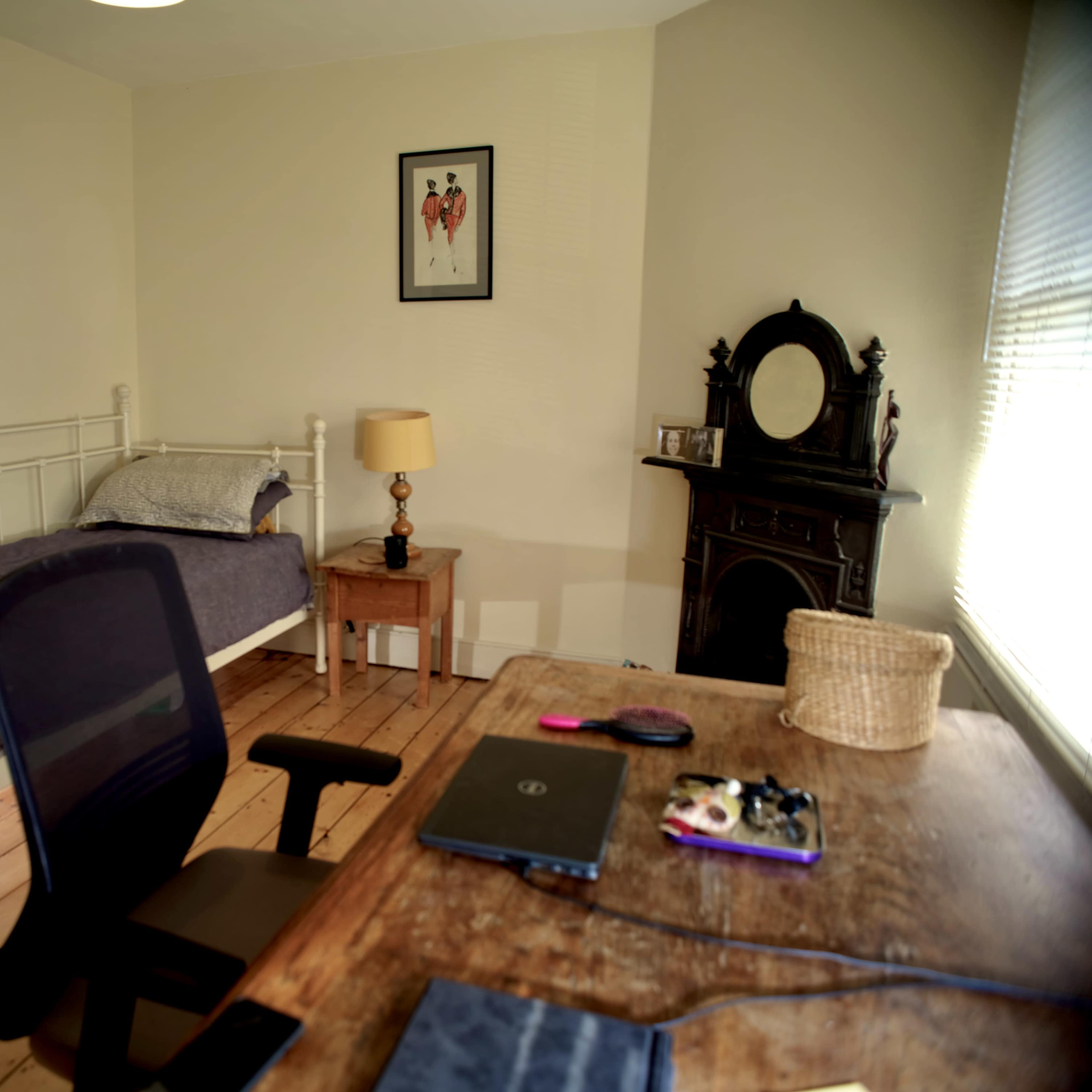 A simple bedroom with a bed, a small lamp on a bedside table, a black chair at a desk, and a decorative mirror on the wall.