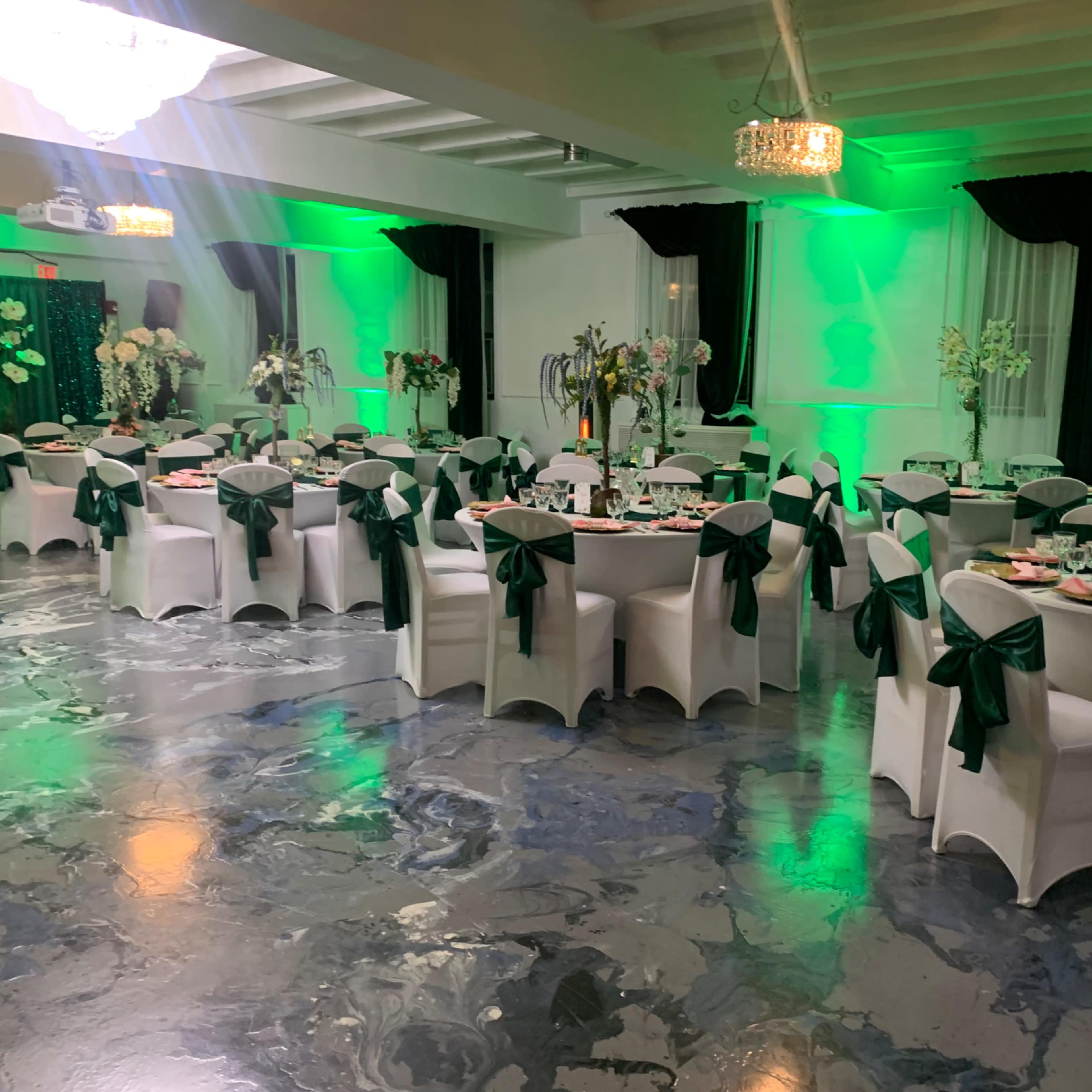A decorated banquet hall with tables set for an event, featuring green-lit walls and centerpieces of flowers.