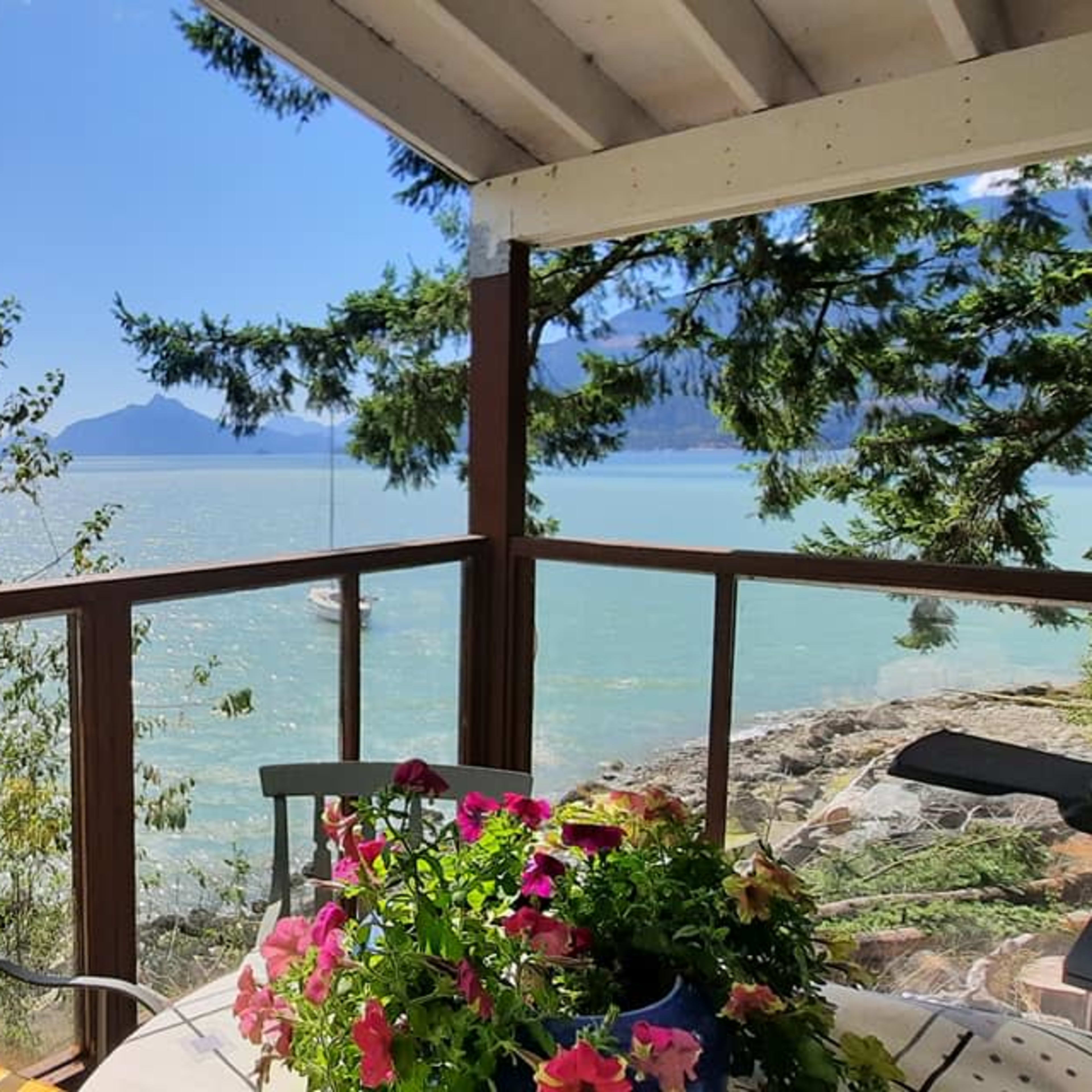A wooden balcony overlooking a calm body of water surrounded by mountains and trees, with a table adorned with flowering plants.