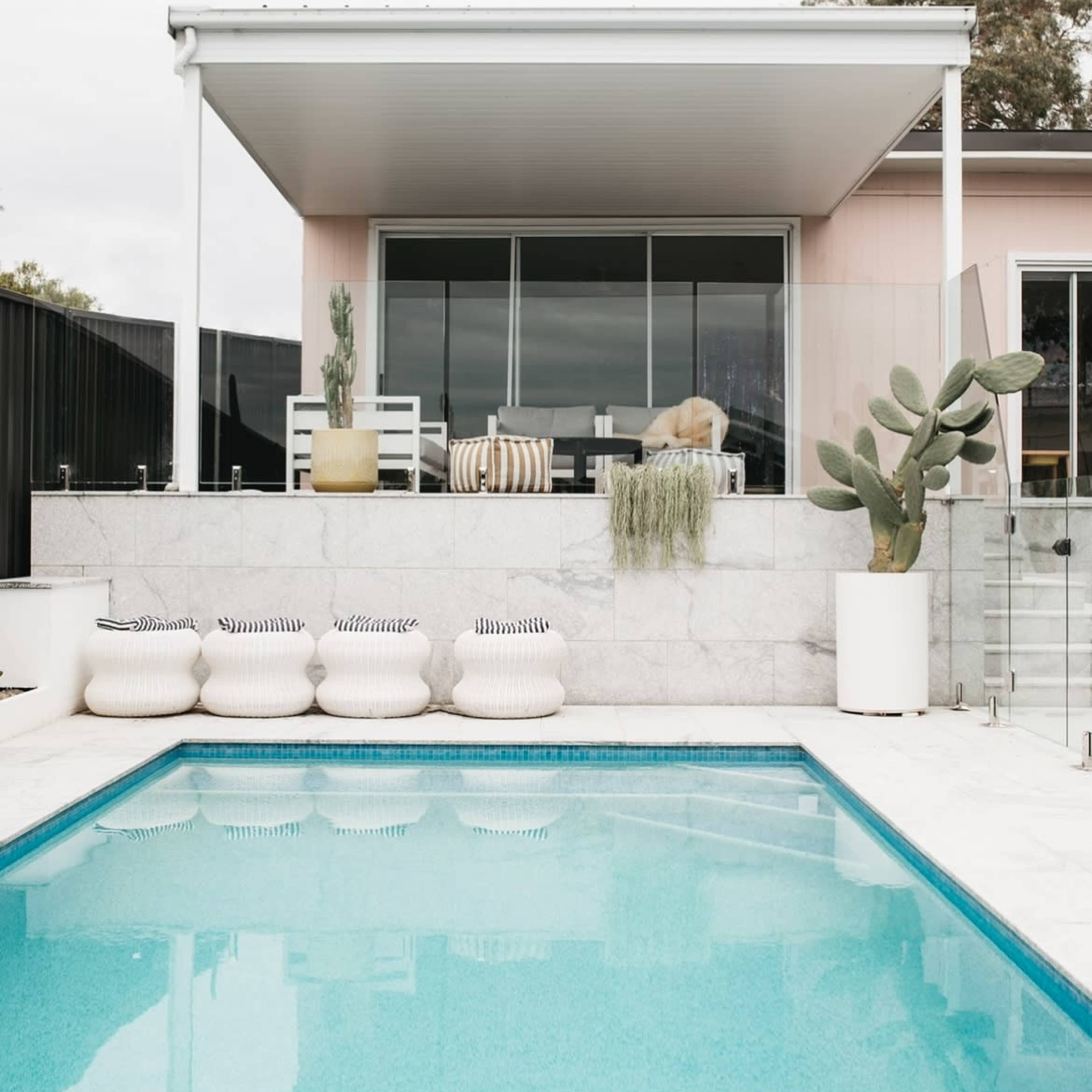 A modern house with a large covered patio overlooks a clear swimming pool, surrounded by potted plants and a stone wall.