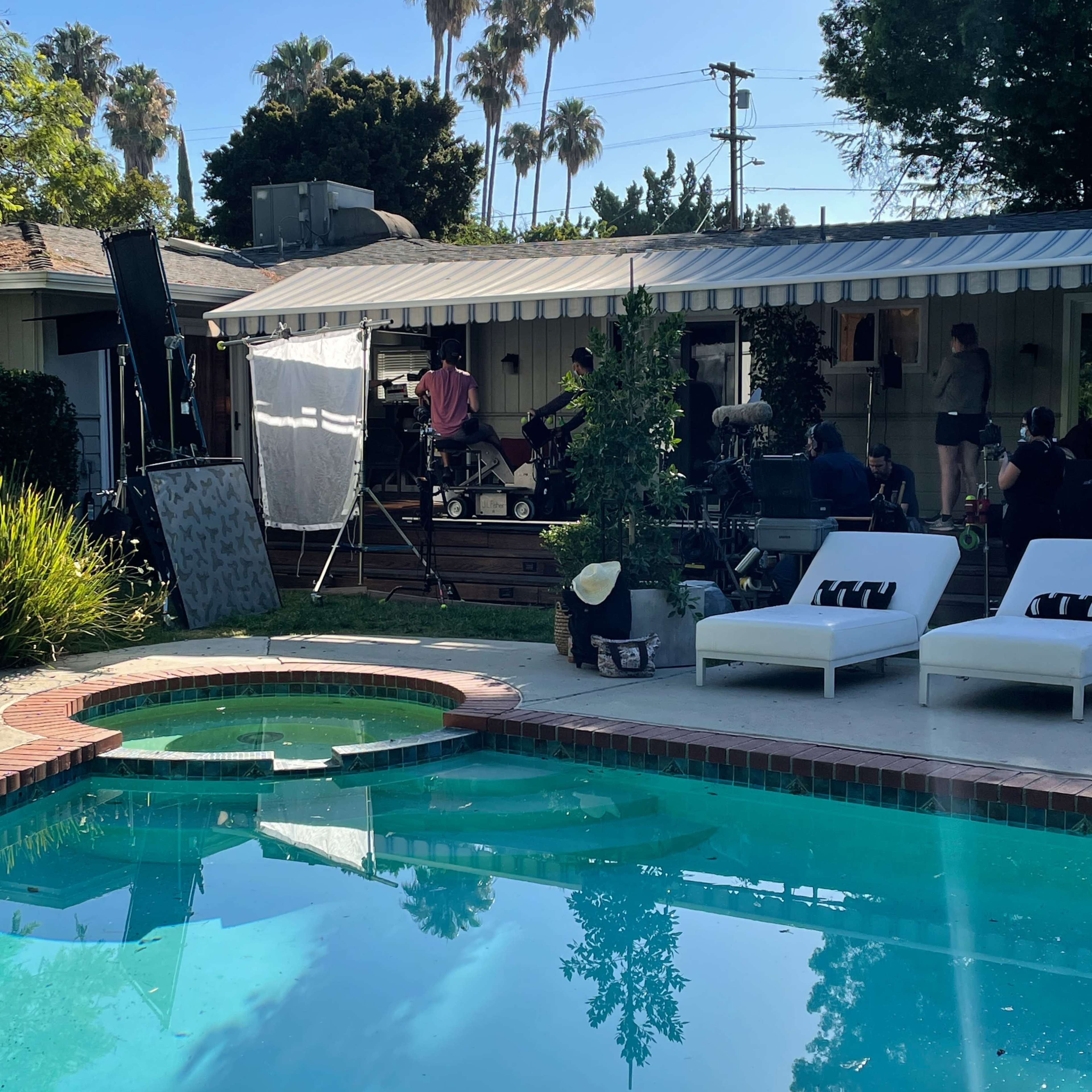 A film crew sets up equipment near a swimming pool surrounded by lounge chairs in a backyard.