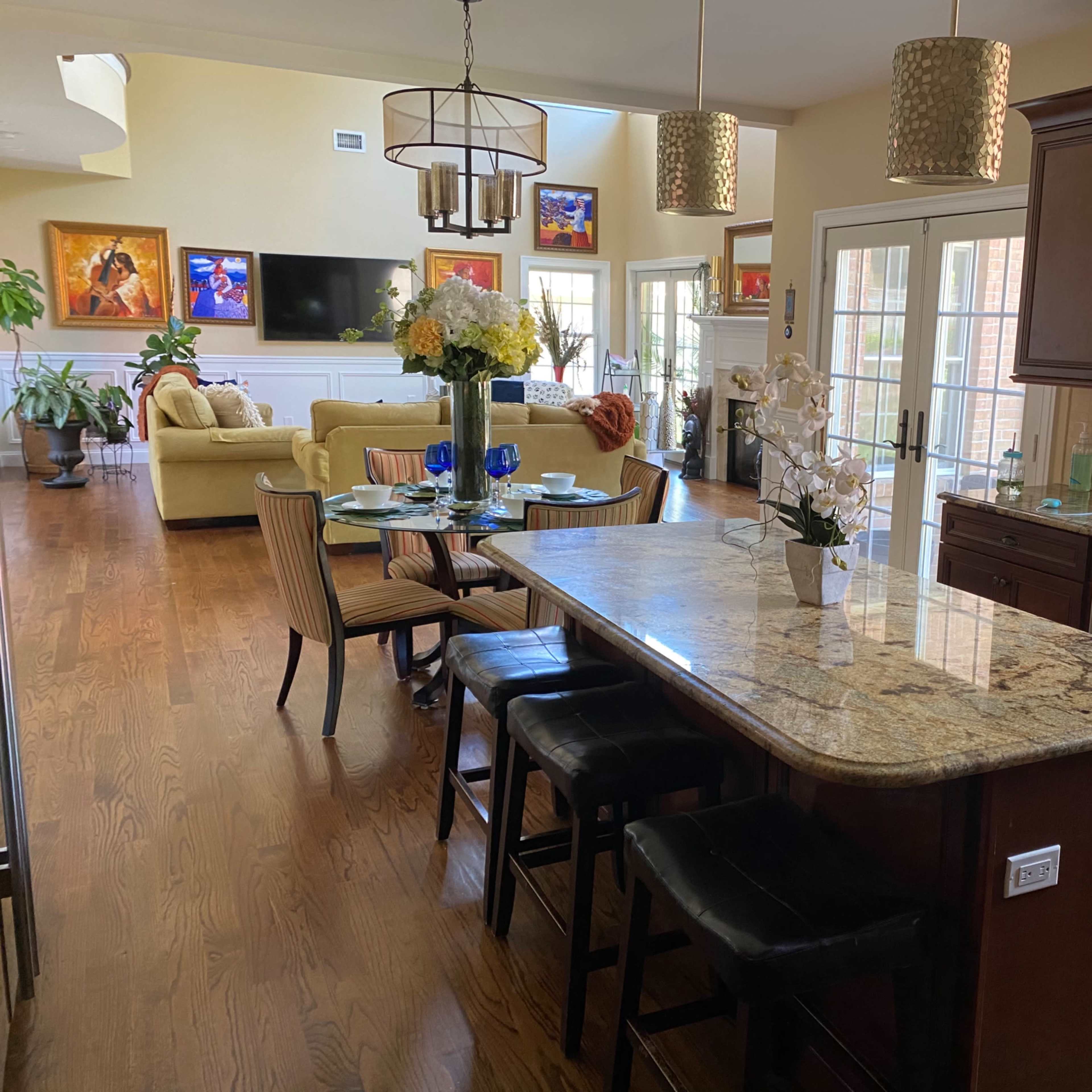 A modern kitchen with a granite island, bar stools, and an open layout leading into a living area with large windows and colorful artwork.