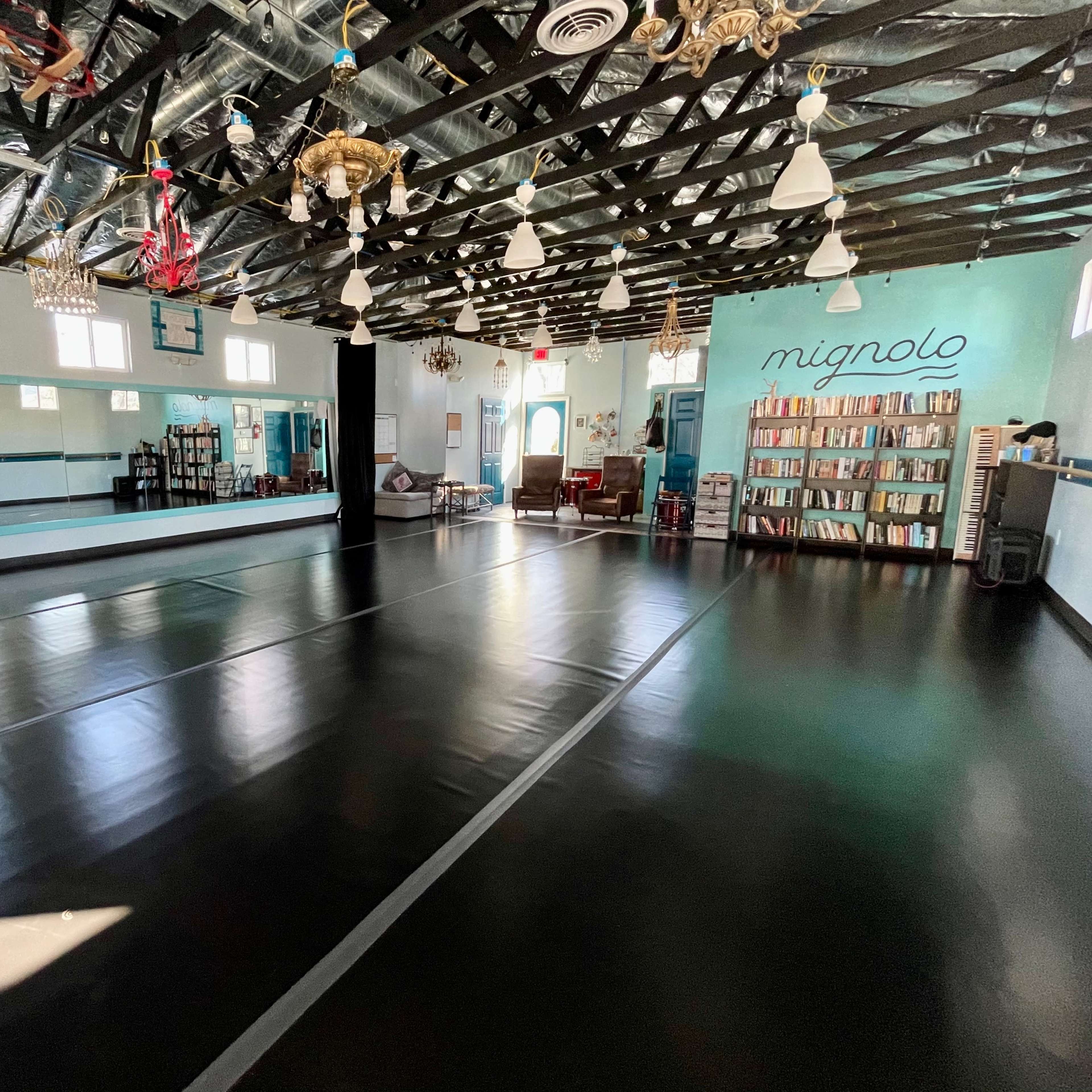 A spacious dance studio with black flooring, large mirrors, and bookshelves along one wall.