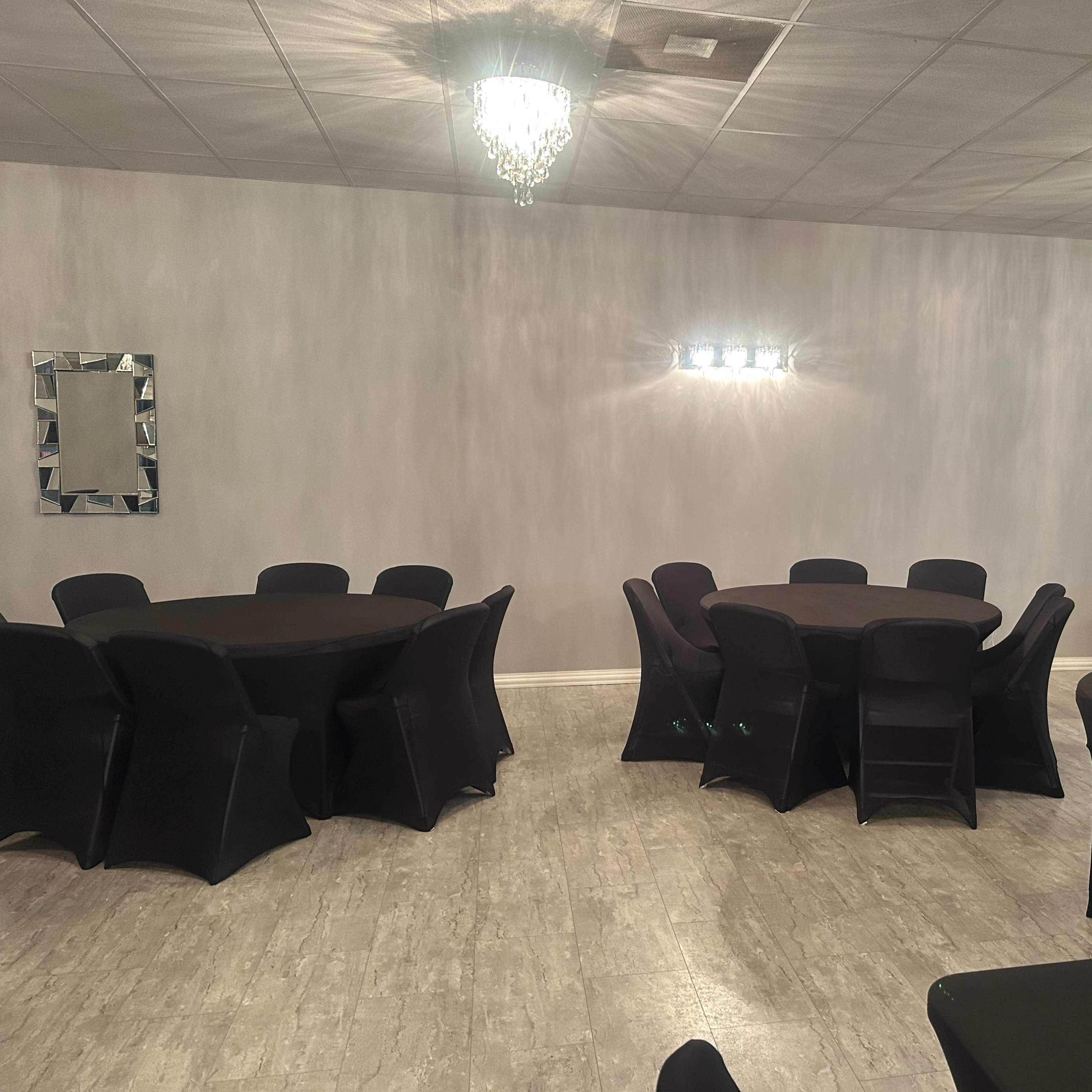 A spacious event room with several round black tables and chairs, illuminated by wall-mounted lights and crystal chandeliers.