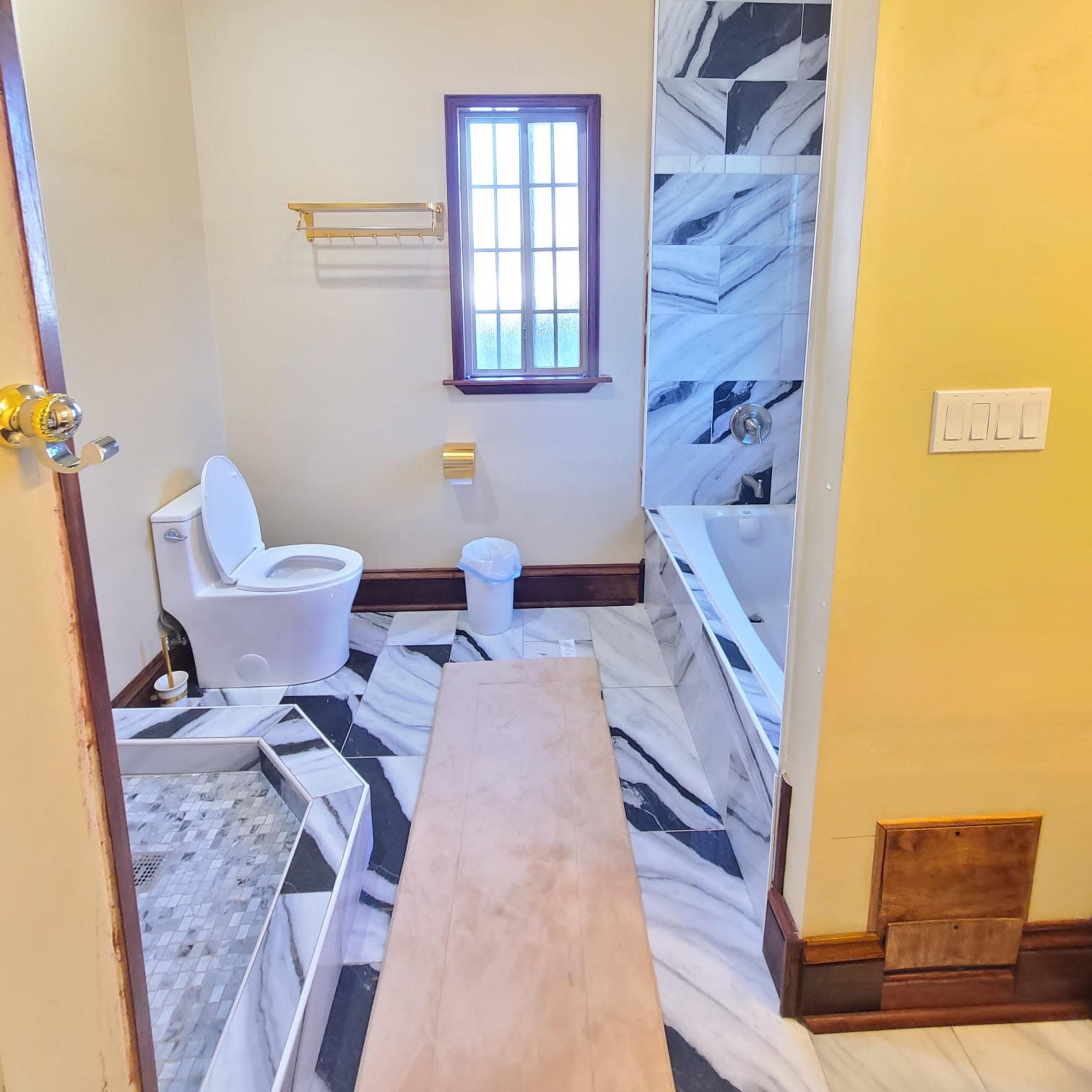 A bathroom with a toilet, bathtub, and a shower area featuring marble flooring and walls, along with a window and some construction materials on the floor.