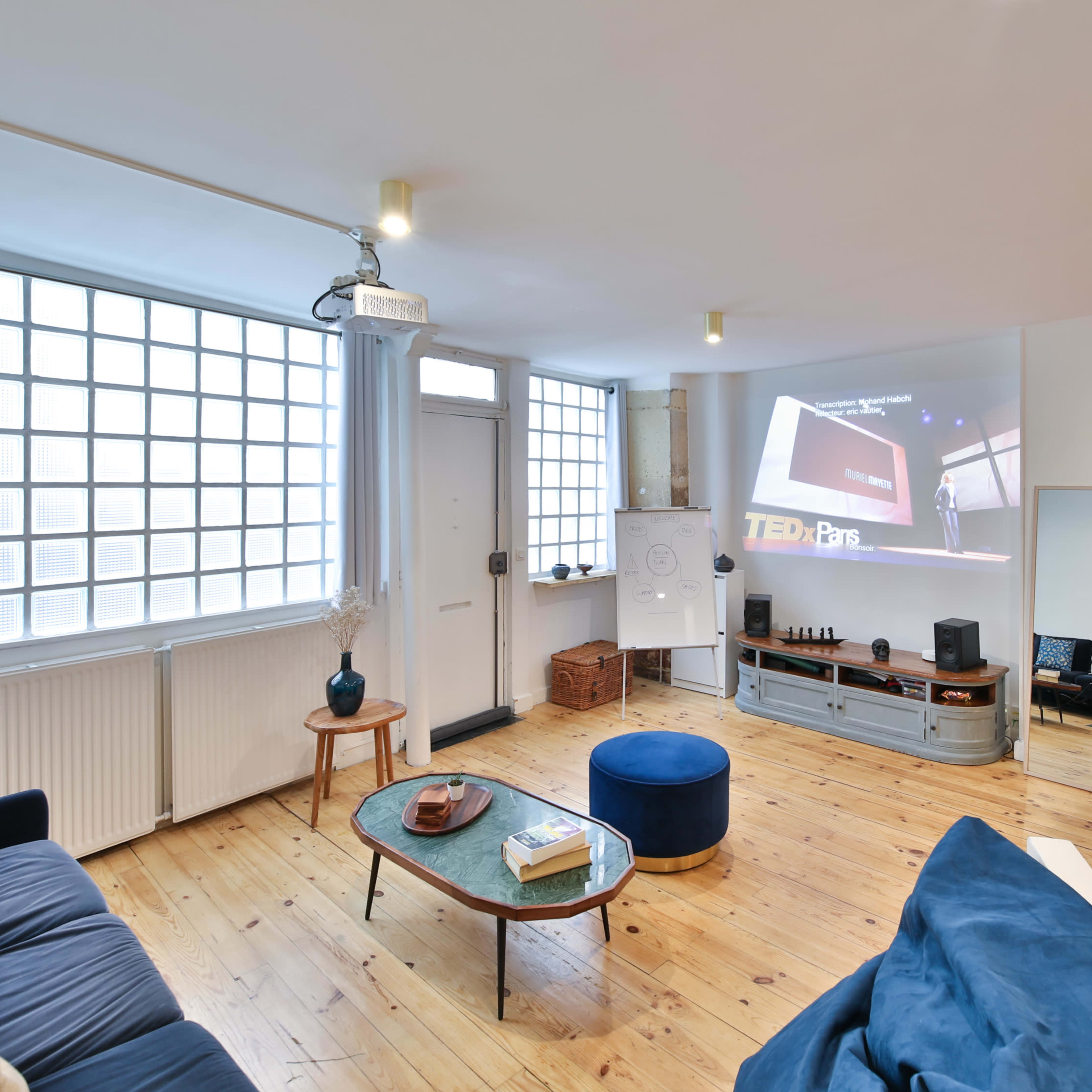 A bright living room with a couch, a coffee table, and a projector displaying a TED talk on the wall.