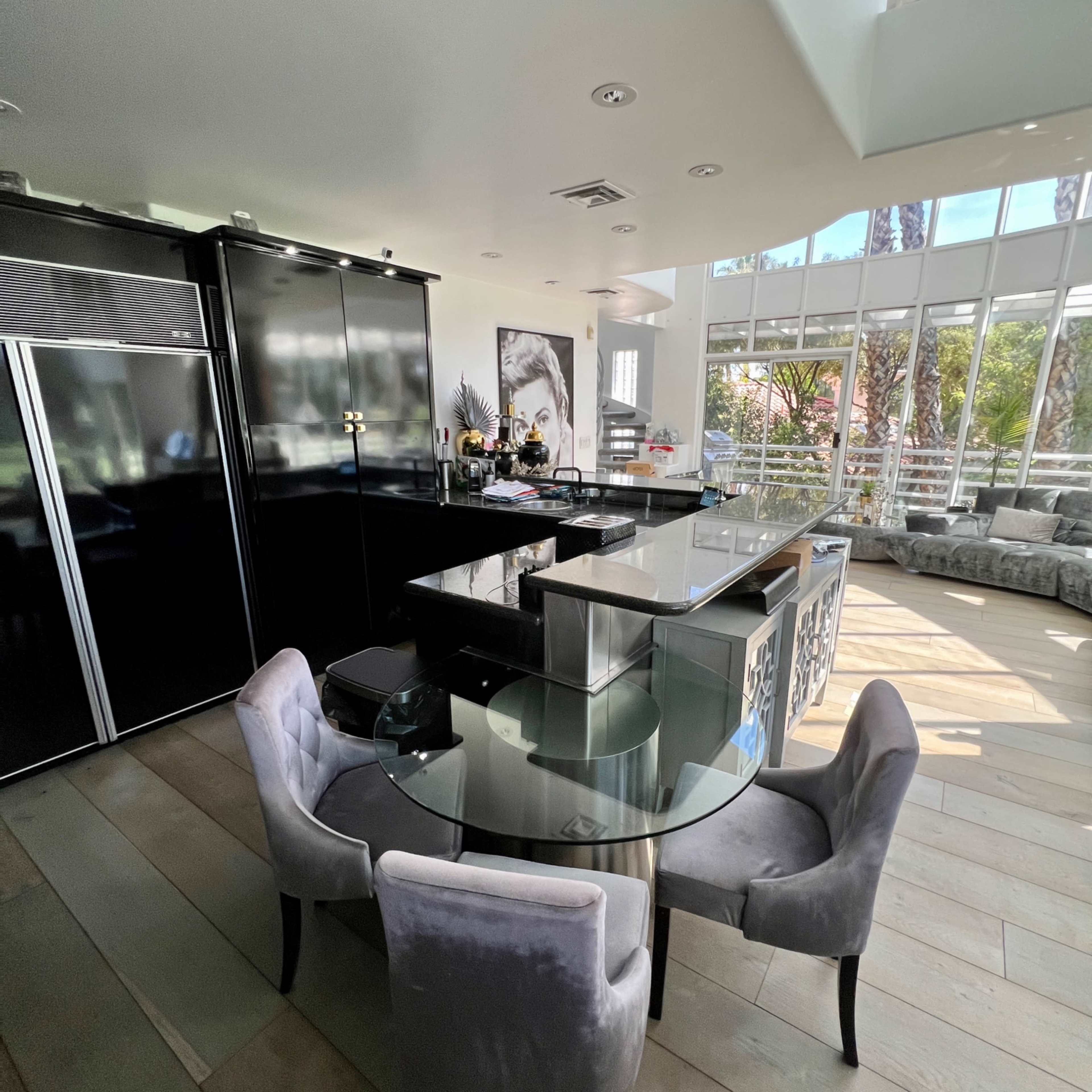 A modern kitchen features a glass dining table surrounded by four plush chairs, with black cabinetry and large windows allowing natural light to fill the space.