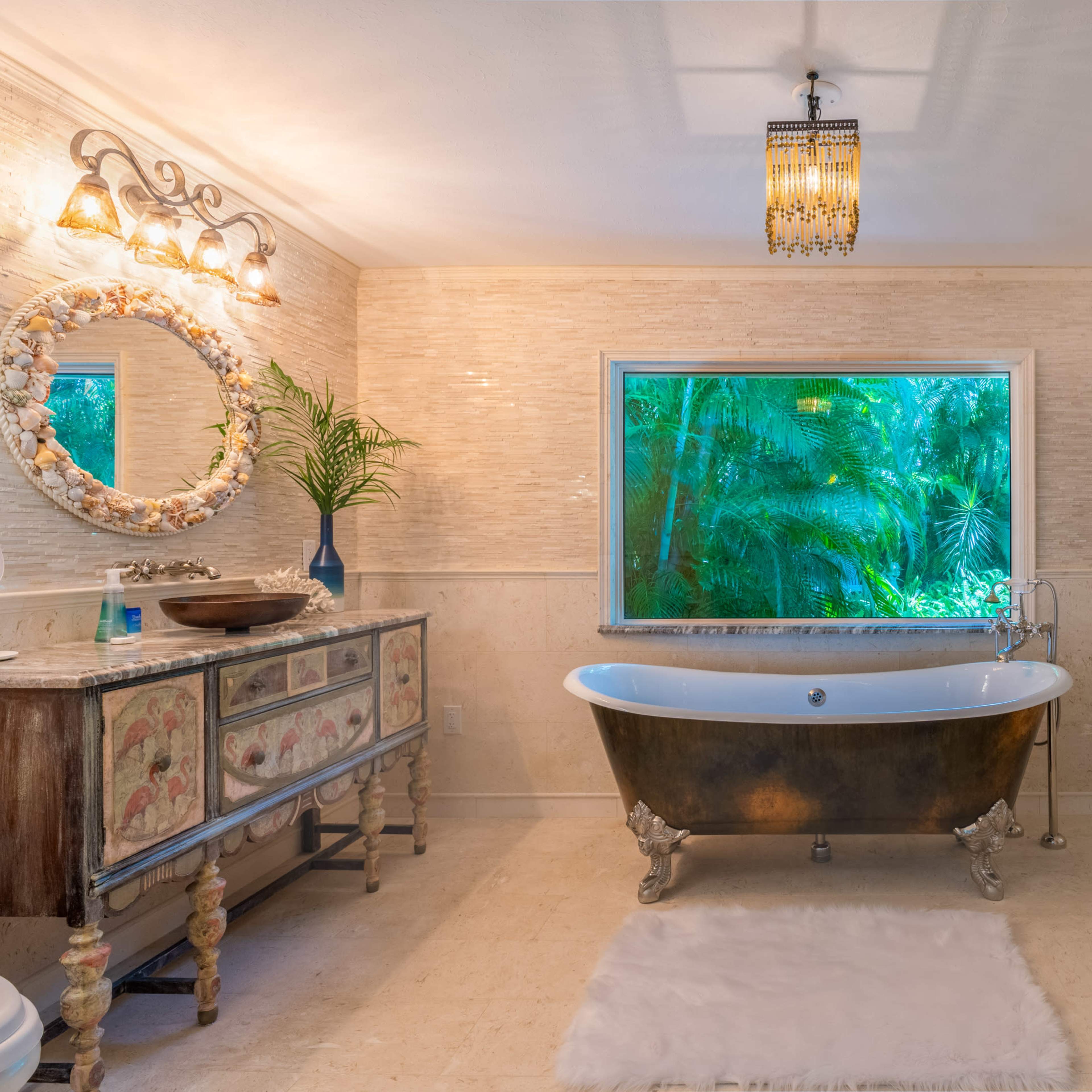 A spacious bathroom features a freestanding black and gold bathtub, a large window framed by greenery, and decorative elements like a vintage mirror and a side cabinet.