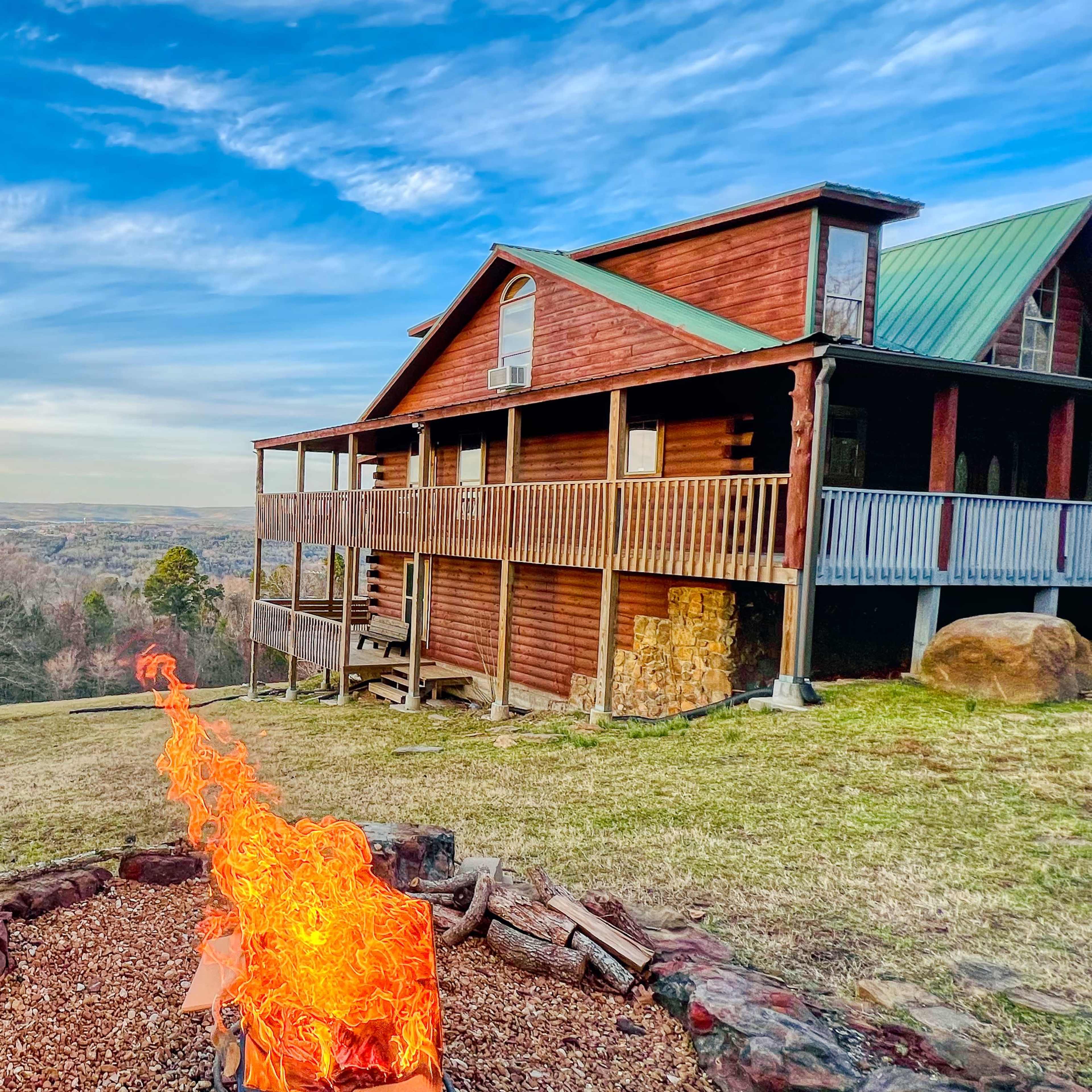 A wooden cabin with a green metal roof overlooks a valley, with a fire pit emitting flames in the foreground.