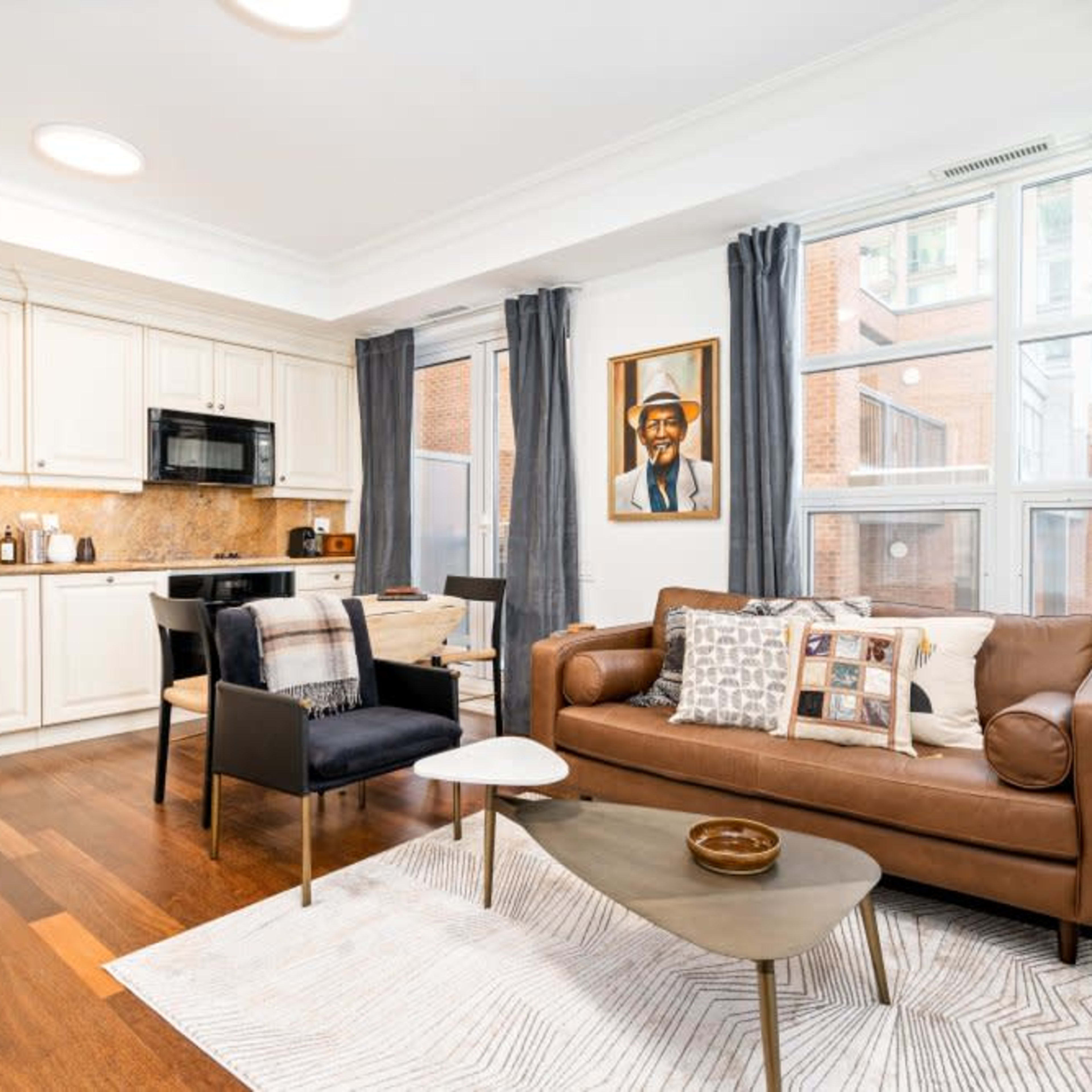 A modern living room with a kitchen area featuring a brown leather sofa, a black armchair, a small coffee table, and large windows letting in natural light.