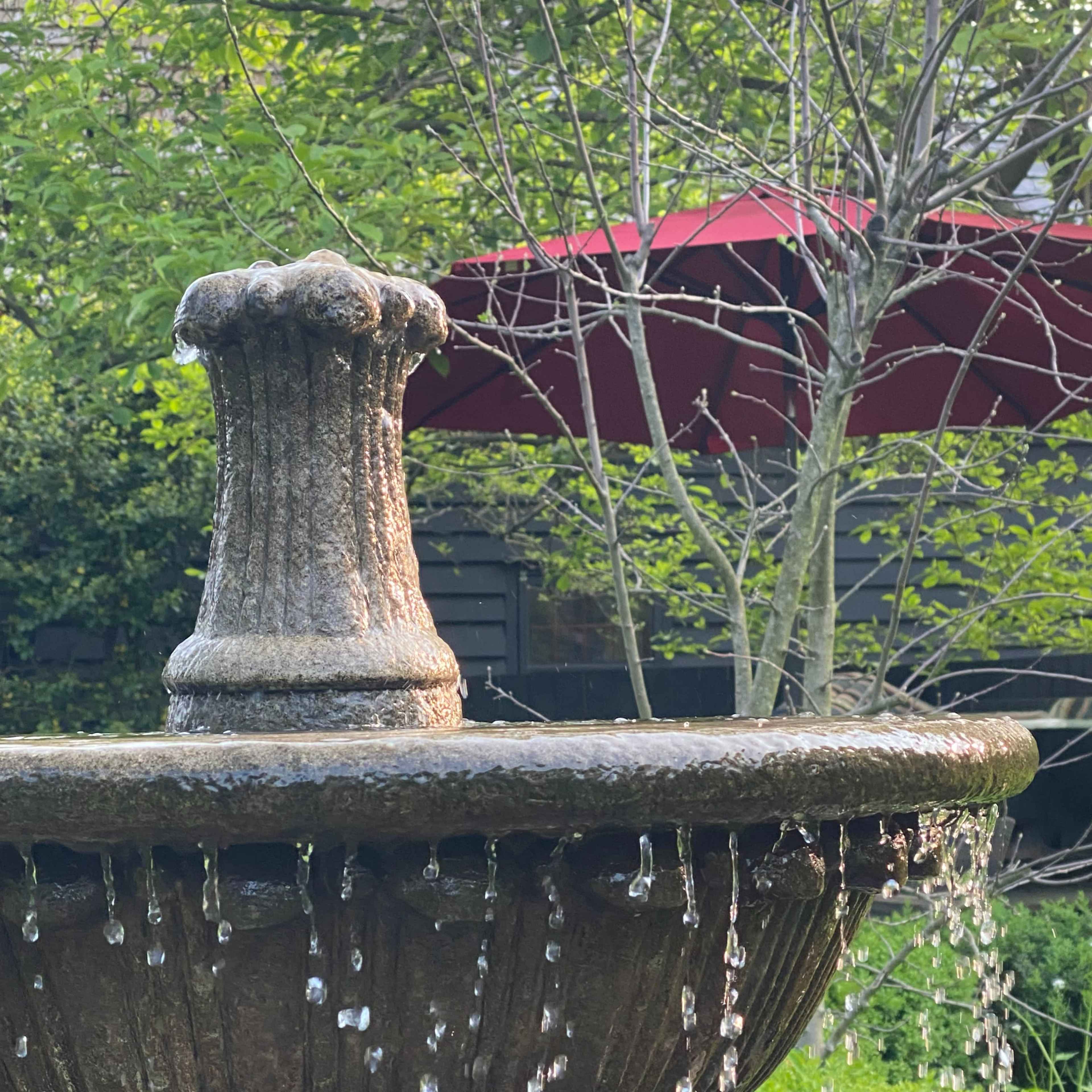 A stone fountain with water cascading from its tiered structure stands in a garden with greenery and a red umbrella in the background.