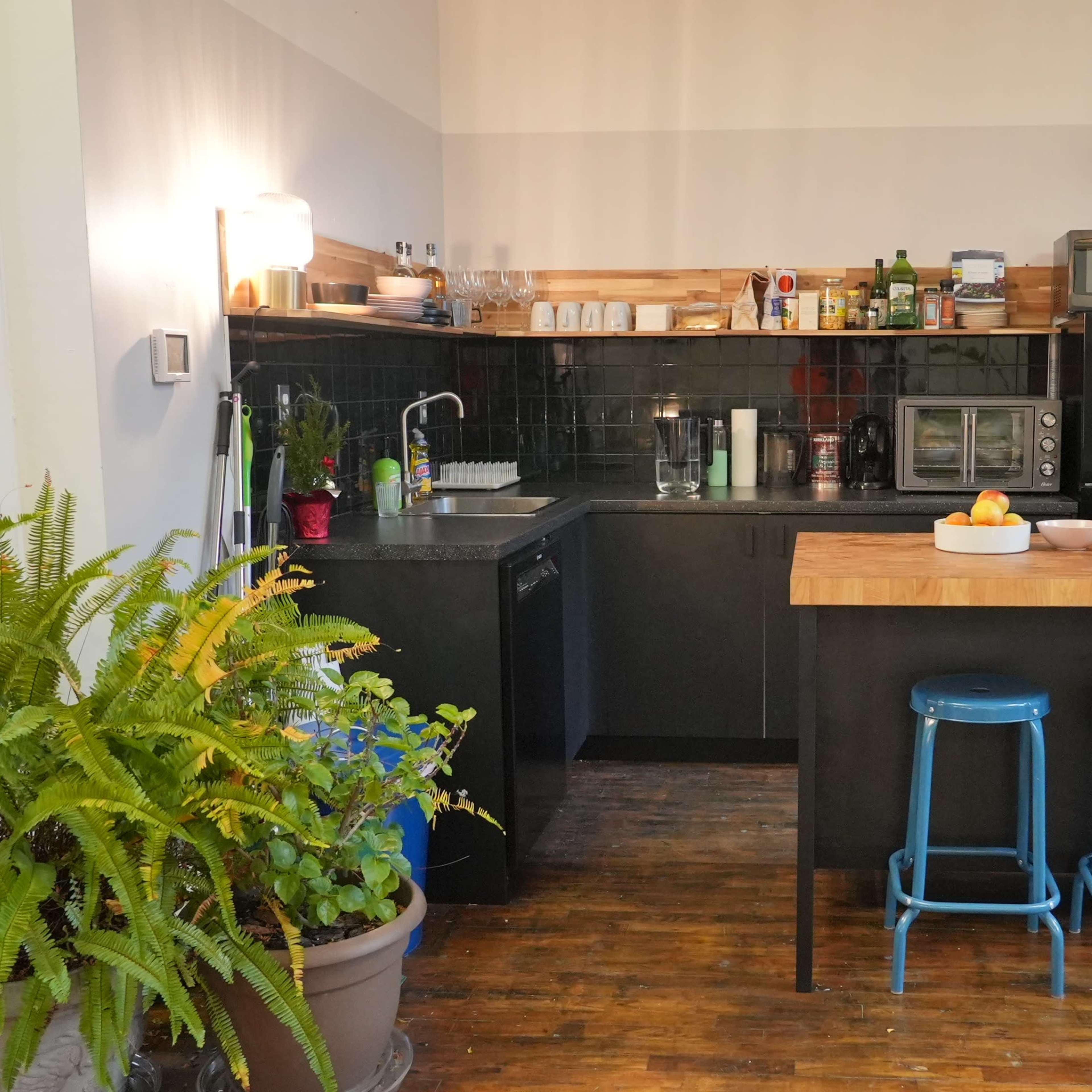 A modern kitchen with a black countertop, a wooden island with blue stools, and potted plants in the background.