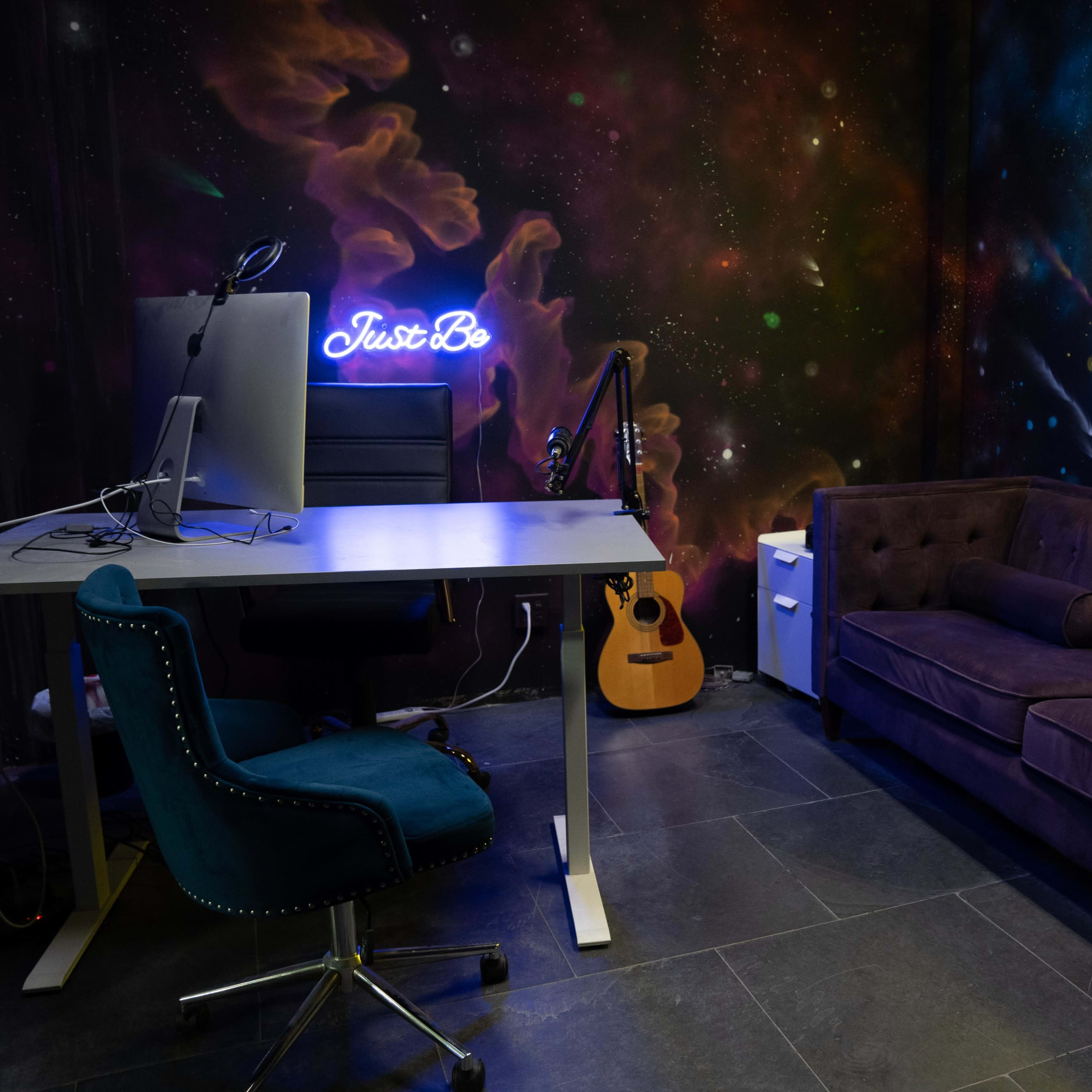 A modern workspace features a desk with a computer, a blue accent chair, a purple sofa, a guitar, and neon signs on a galaxy-themed wall.