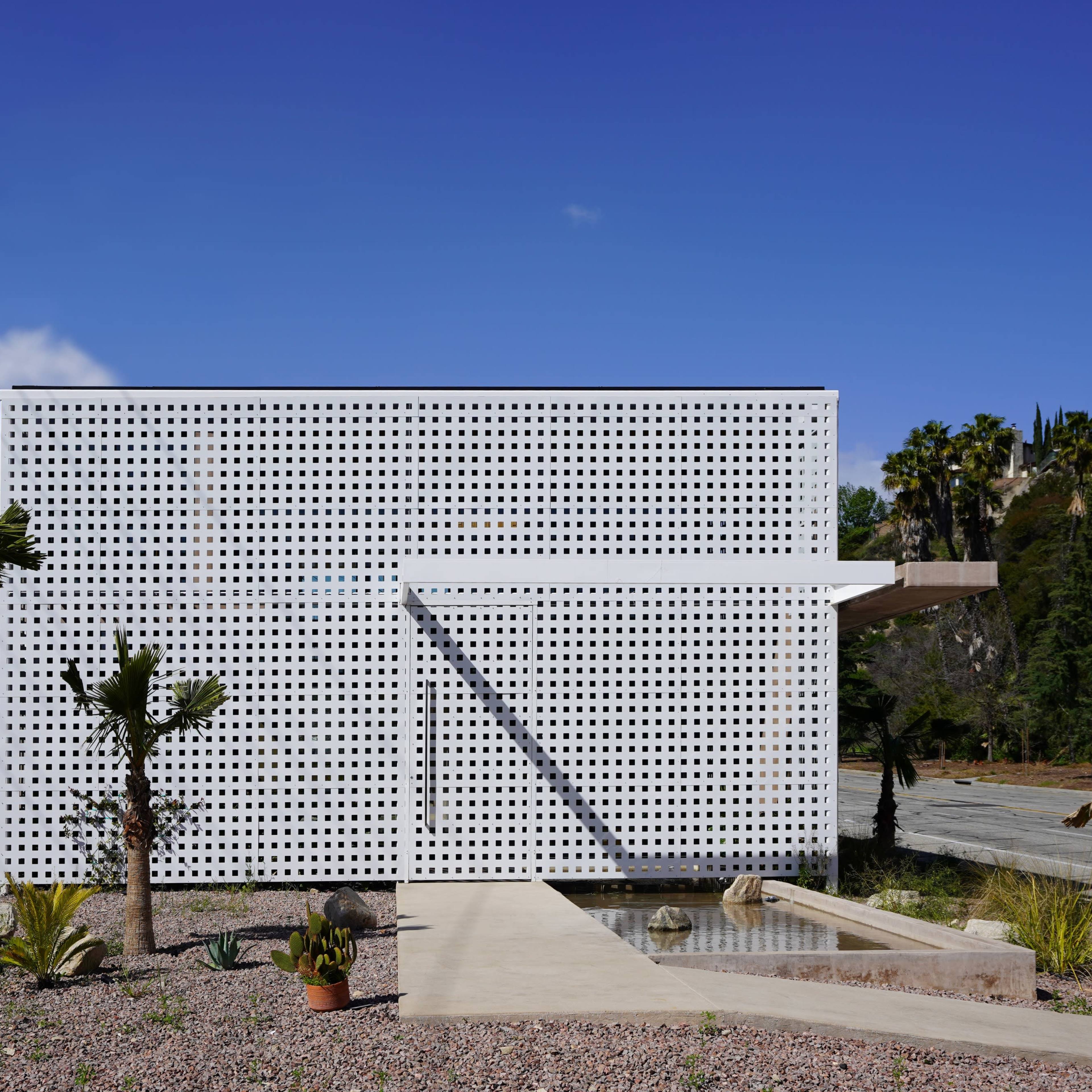 A modern white building with a perforated facade sits in a landscaped area featuring desert plants and a pathway.