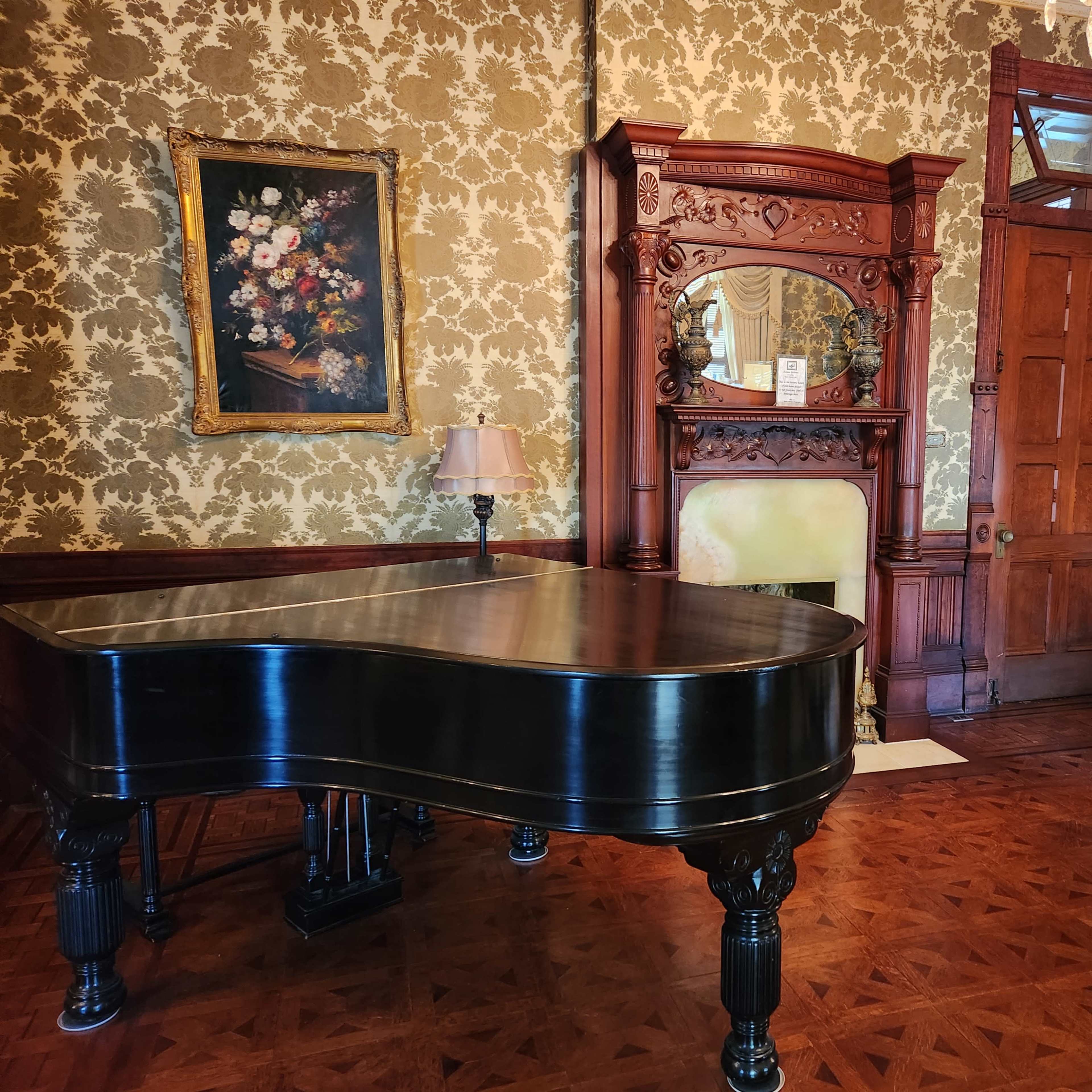 A grand piano sits in a richly decorated room featuring floral wallpaper, a vintage fireplace, and ornate wooden furnishings.