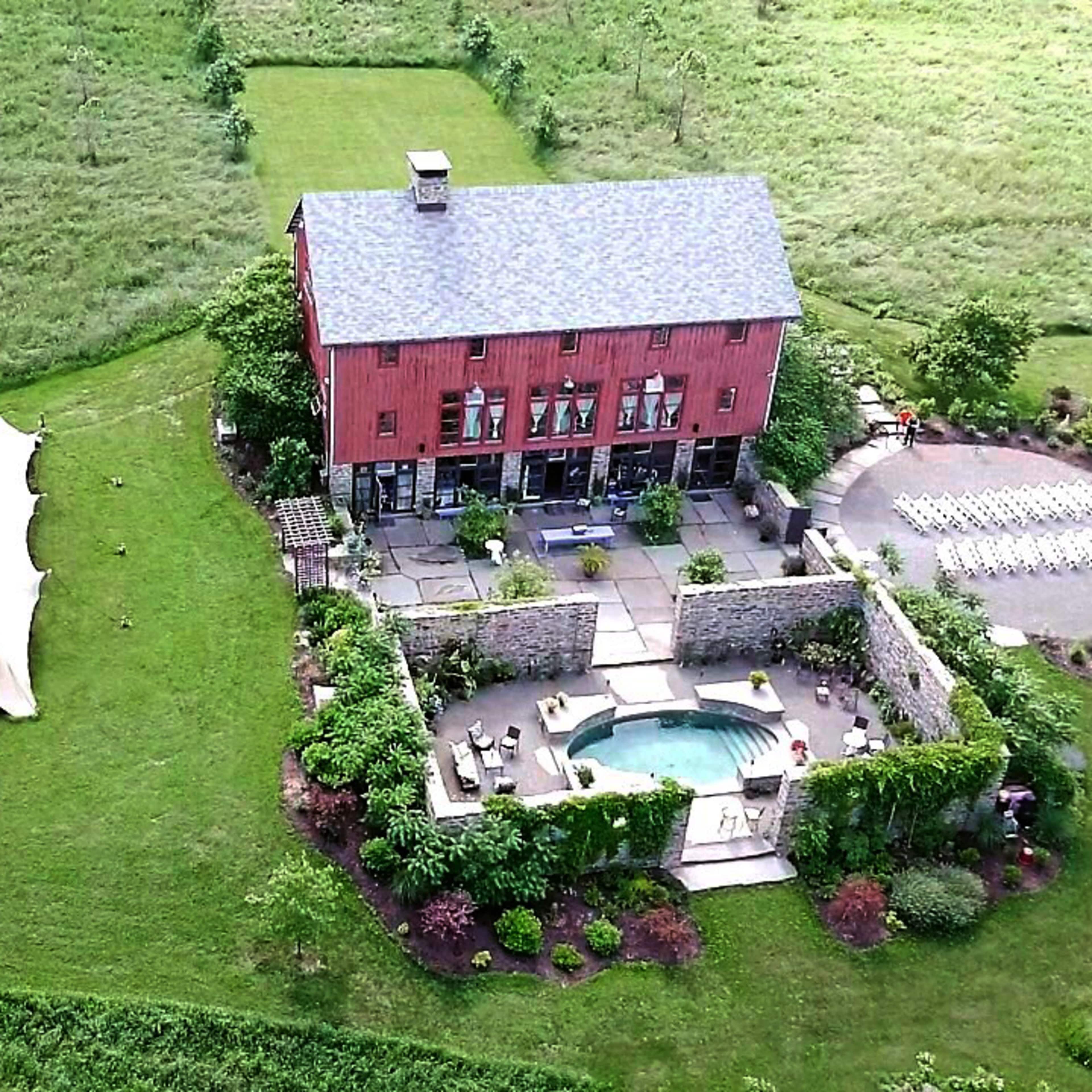 An aerial view shows a large red barn house surrounded by landscaped gardens, a pool, and a tented area on a grassy field.
