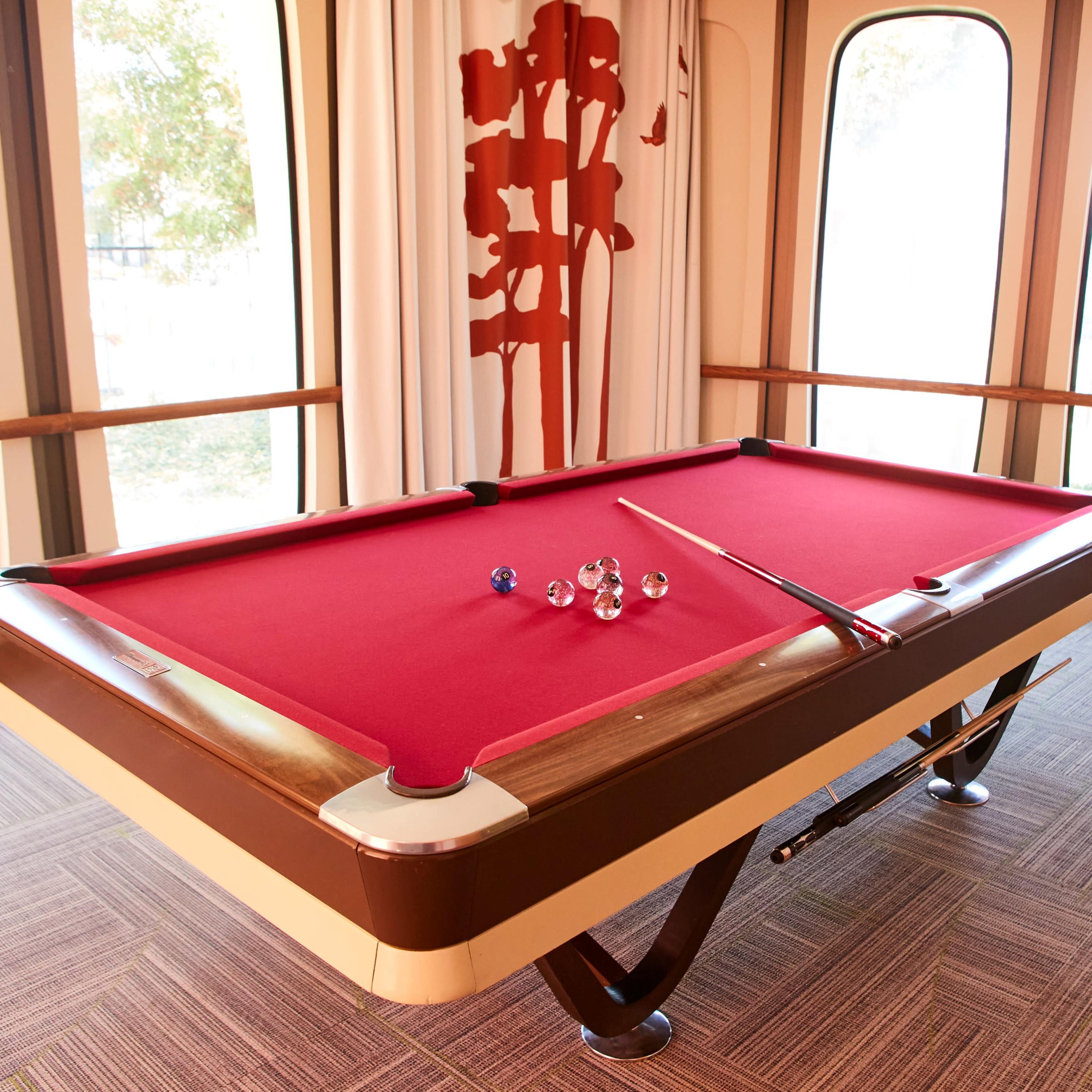 A pool table with a red felt surface and five balls is positioned in a brightly lit room with large windows and decorative curtains.