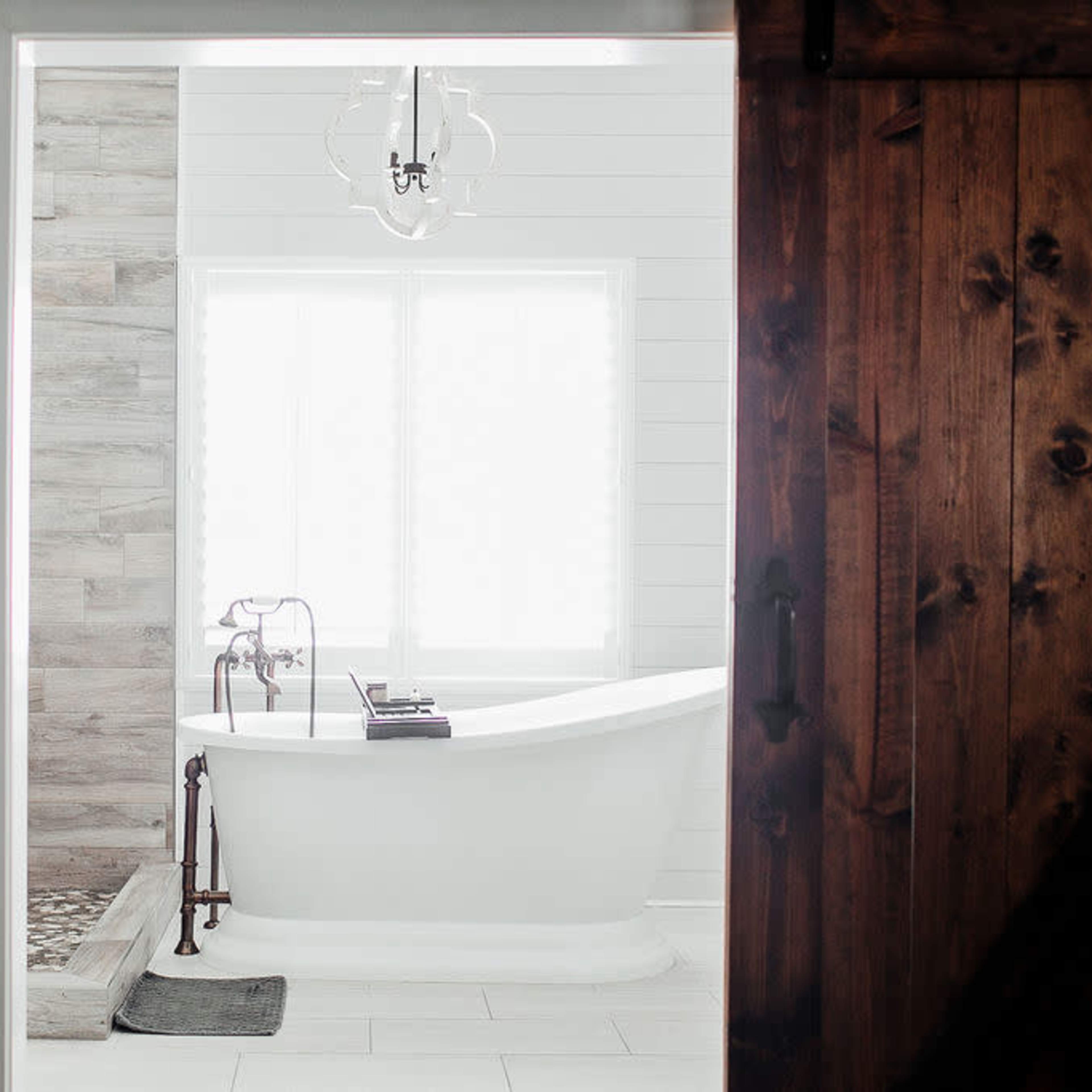 A spacious bathroom featuring a white freestanding bathtub next to a wooden accent wall, with a large window allowing natural light to enter.