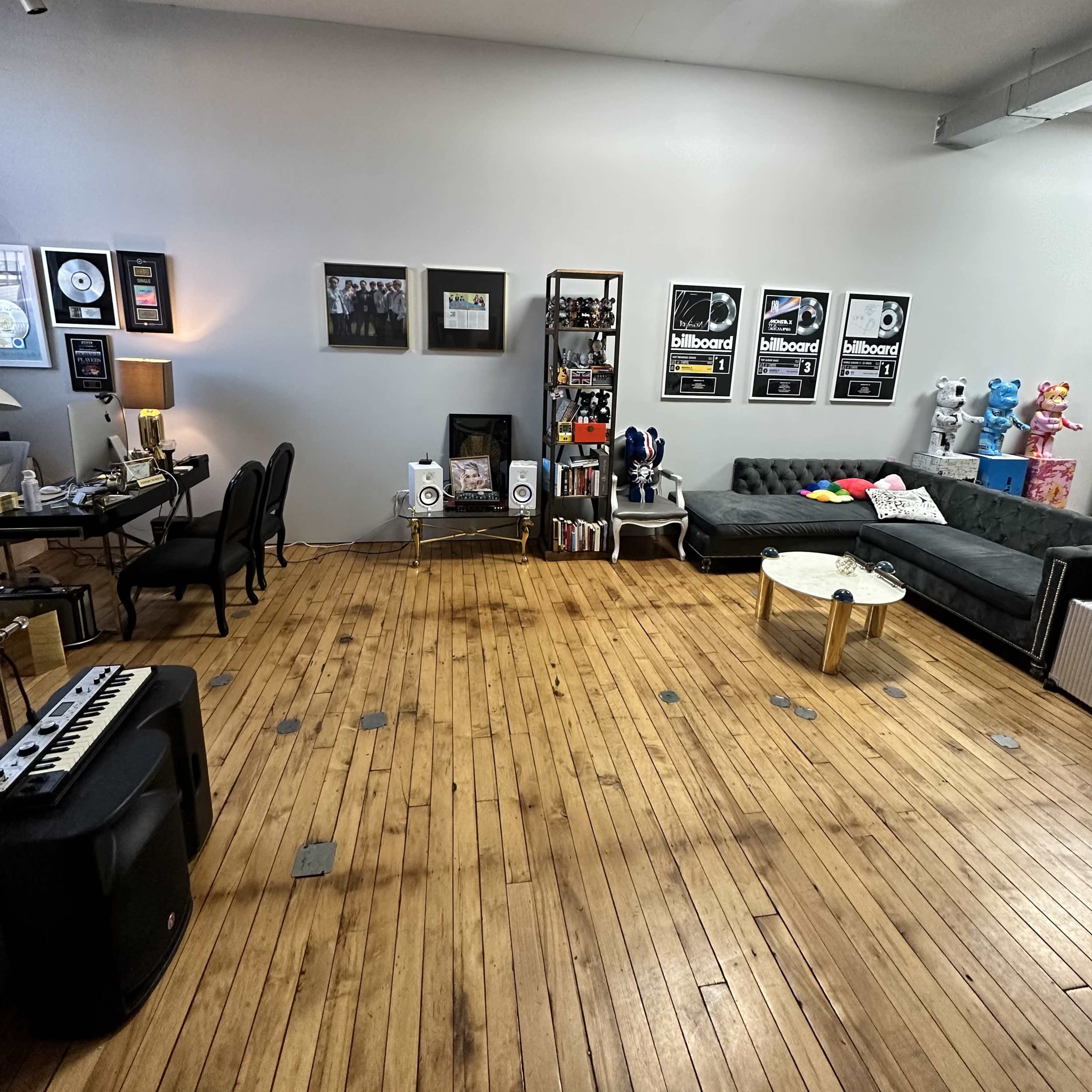 A spacious room with wooden floors, featuring a seating area with a couch and colorful decorations, along with a workspace that includes a desk and musical equipment.