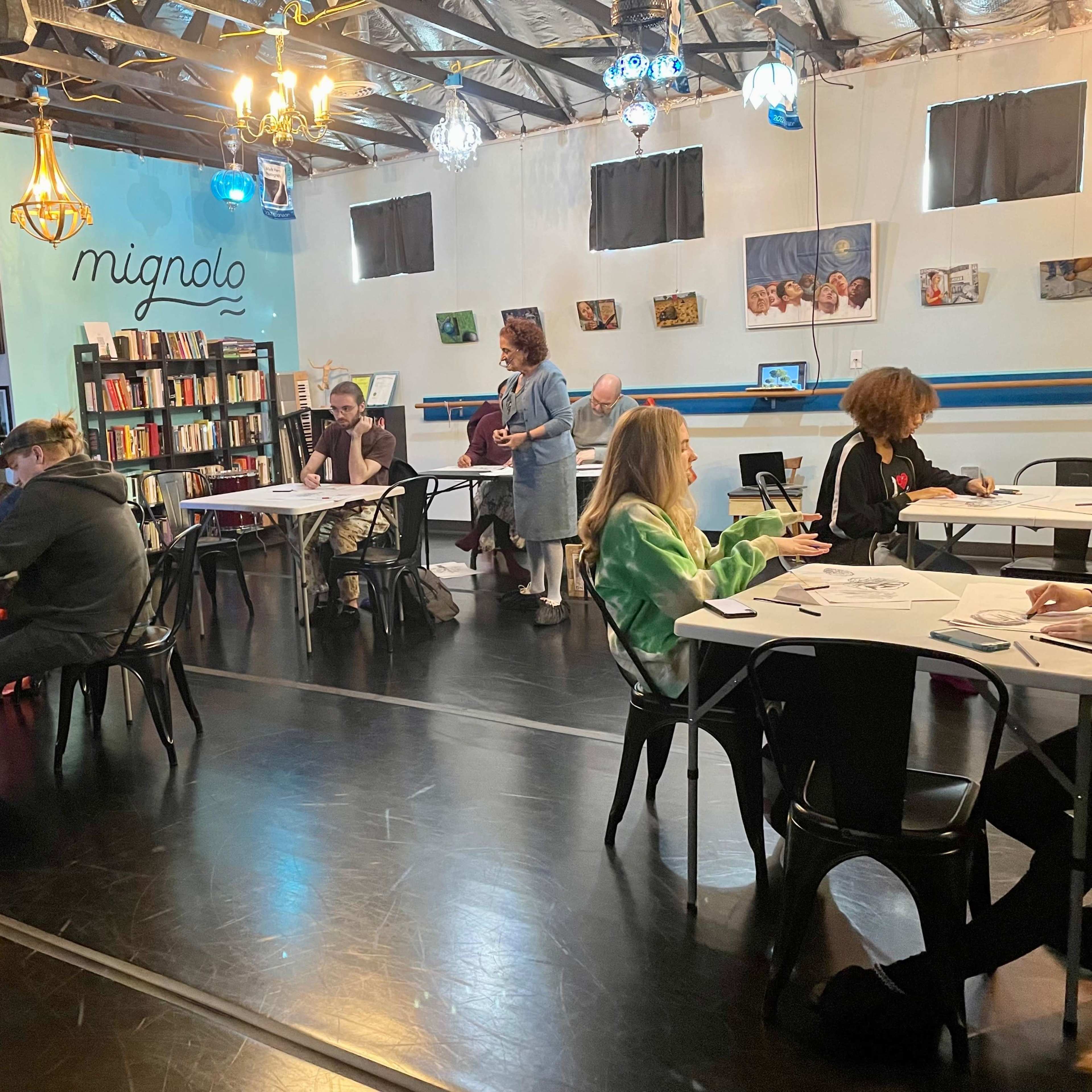 A group of people sits at tables in a brightly lit room with shelves of books and artwork on the walls.