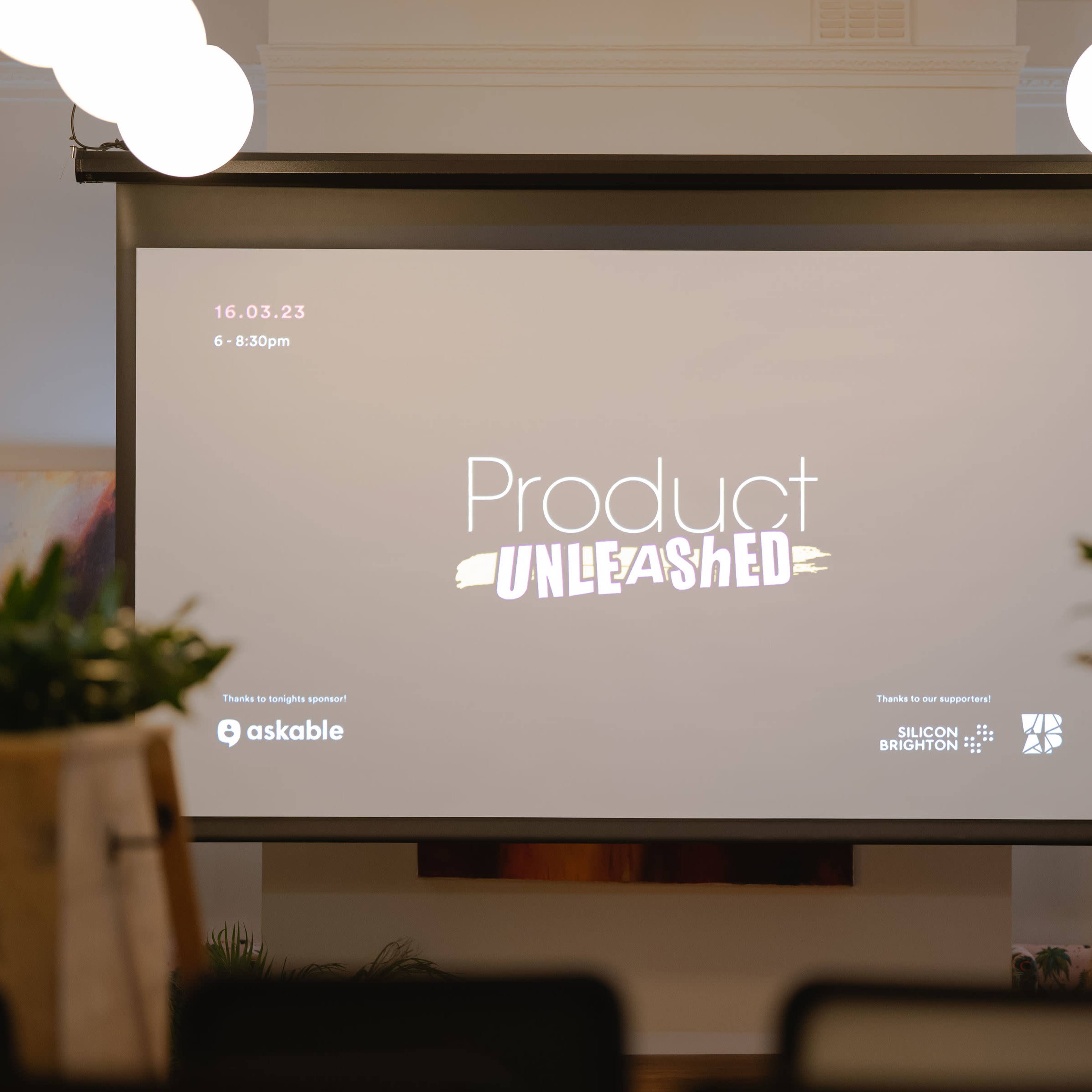 A digital screen displays the event title "Product Unleashed" with the date and time in a venue set for a presentation.