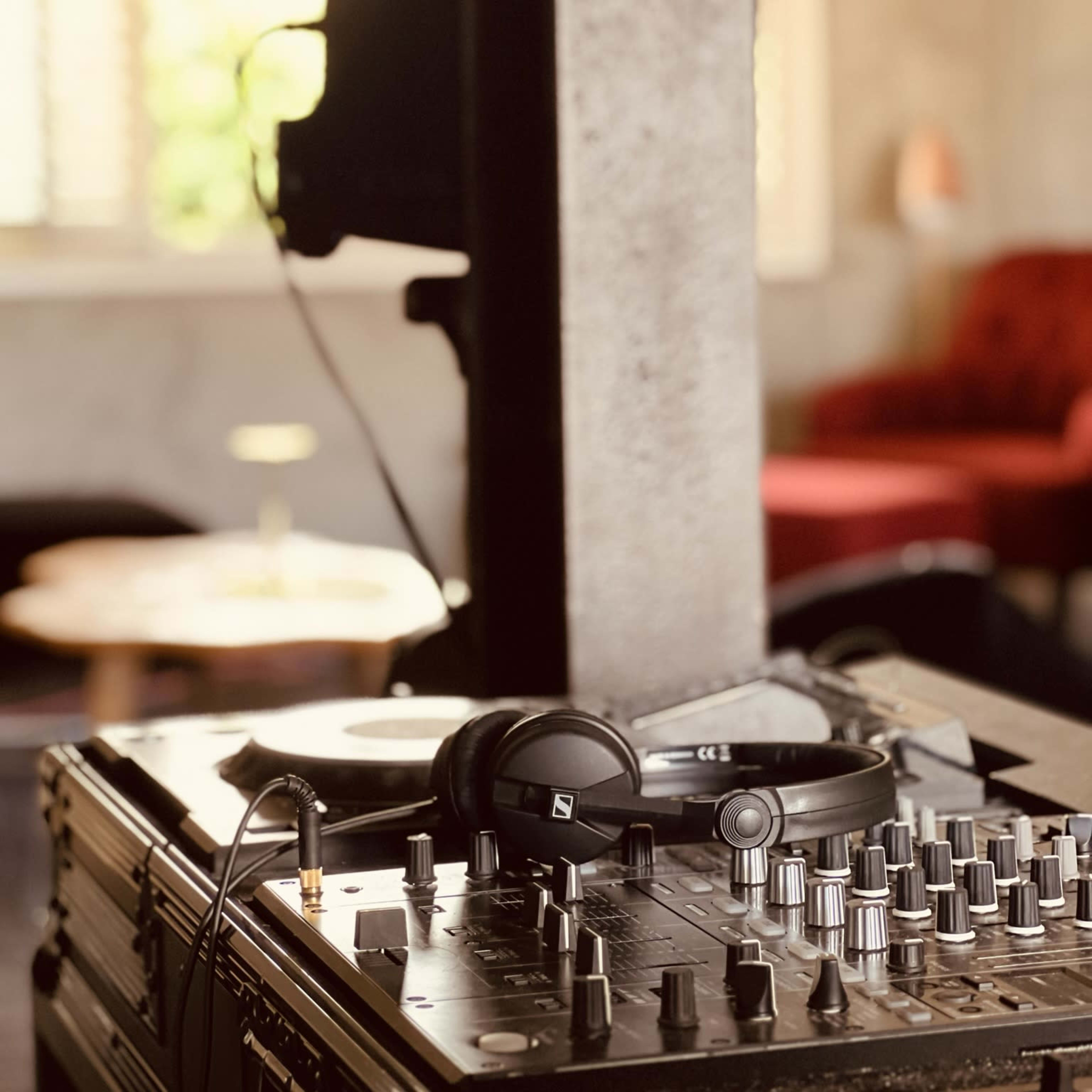 A DJ mixer and headphones are set on a table in a room with a large window and a red chair in the background.