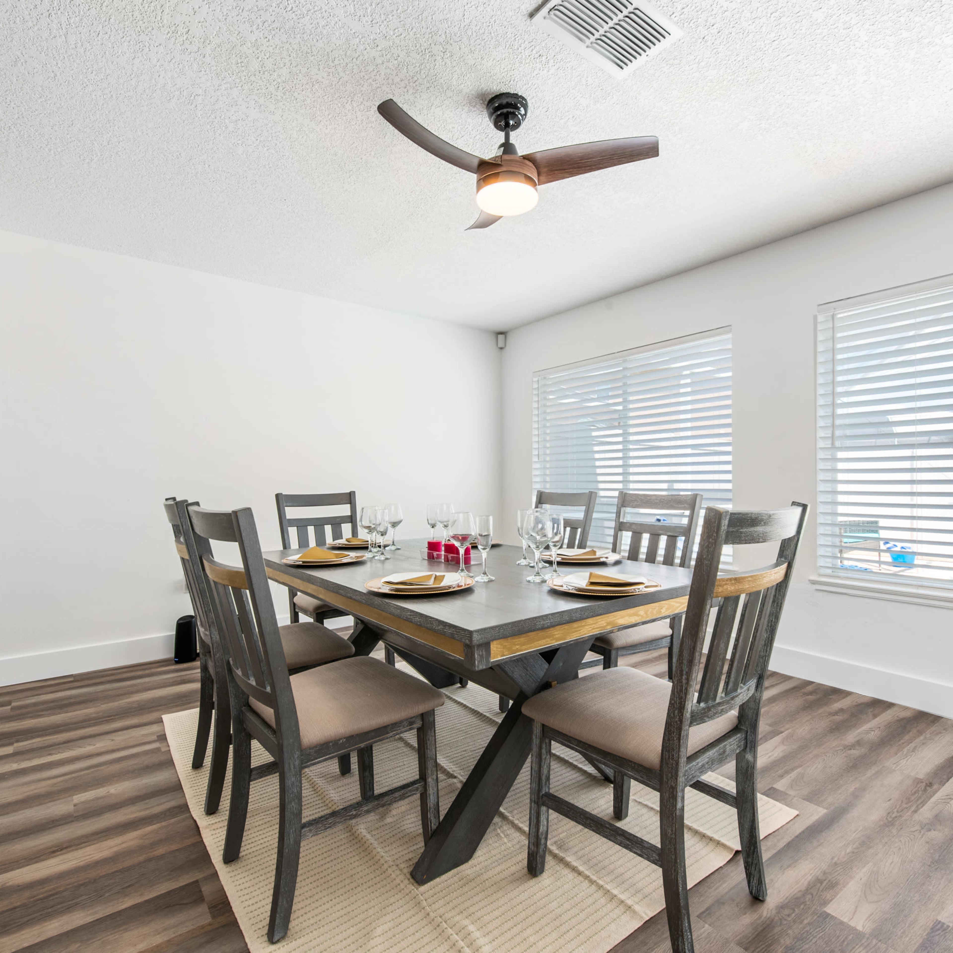 A dining room features a wooden table set for six, surrounded by gray chairs, with ceiling fans and large windows allowing ample natural light.