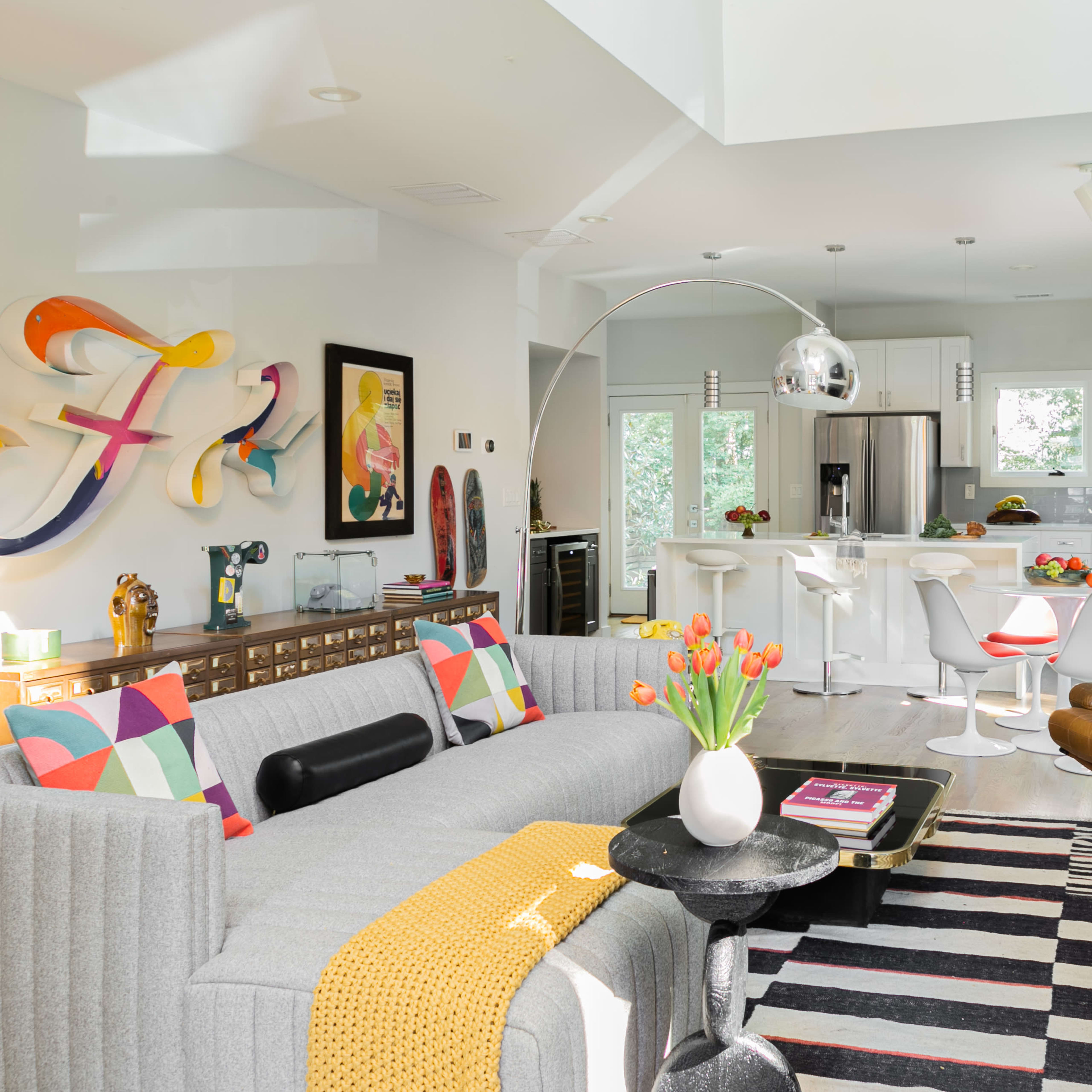 A modern living space features a gray sofa, a black coffee table, colorful decorative pillows, and an open kitchen with white cabinetry and bar stools.